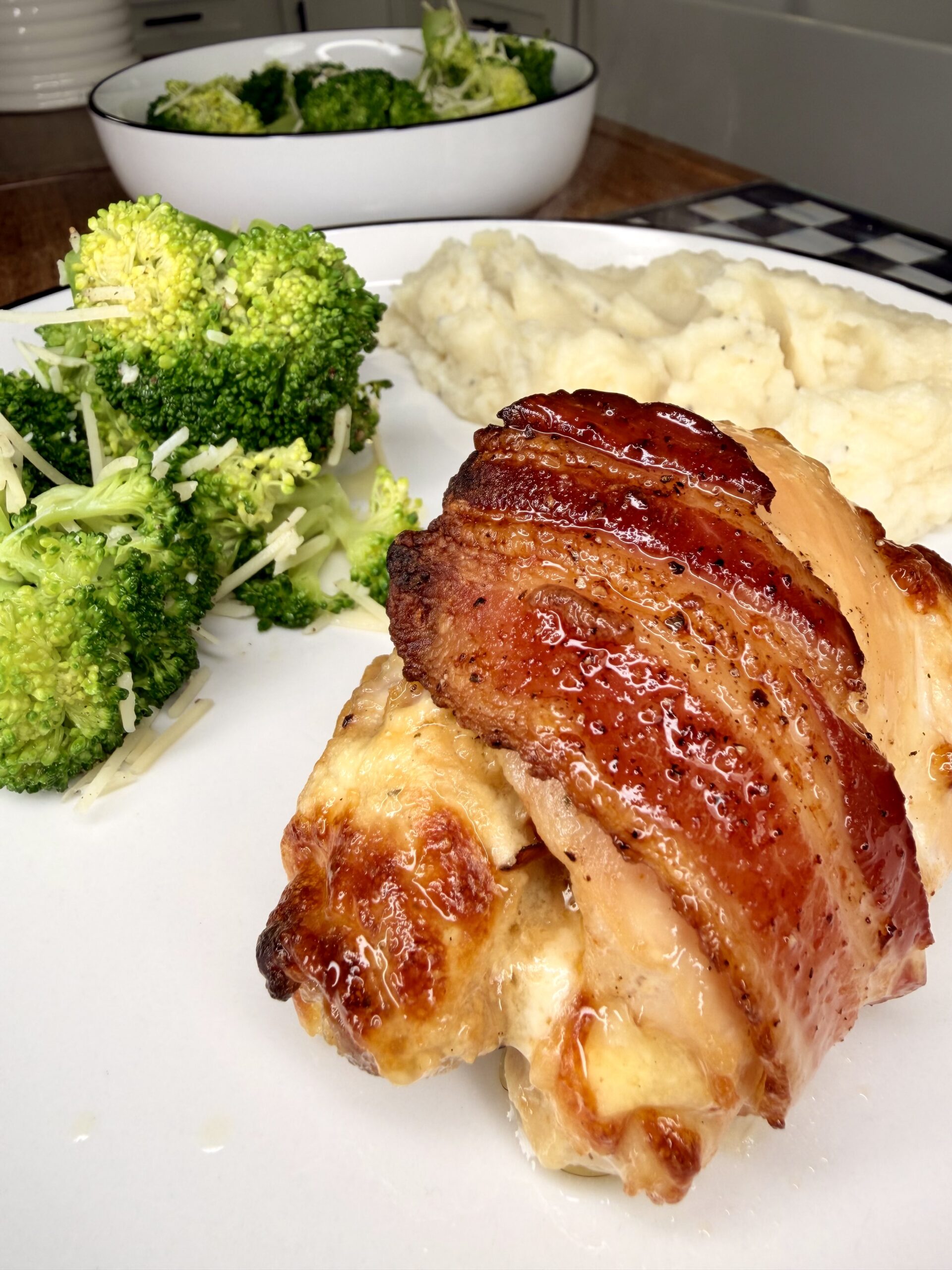 A plate of bacon-wrapped chicken breast, mashed potatoes, and steamed broccoli, with an additional bowl of broccoli in the background on a wooden table.