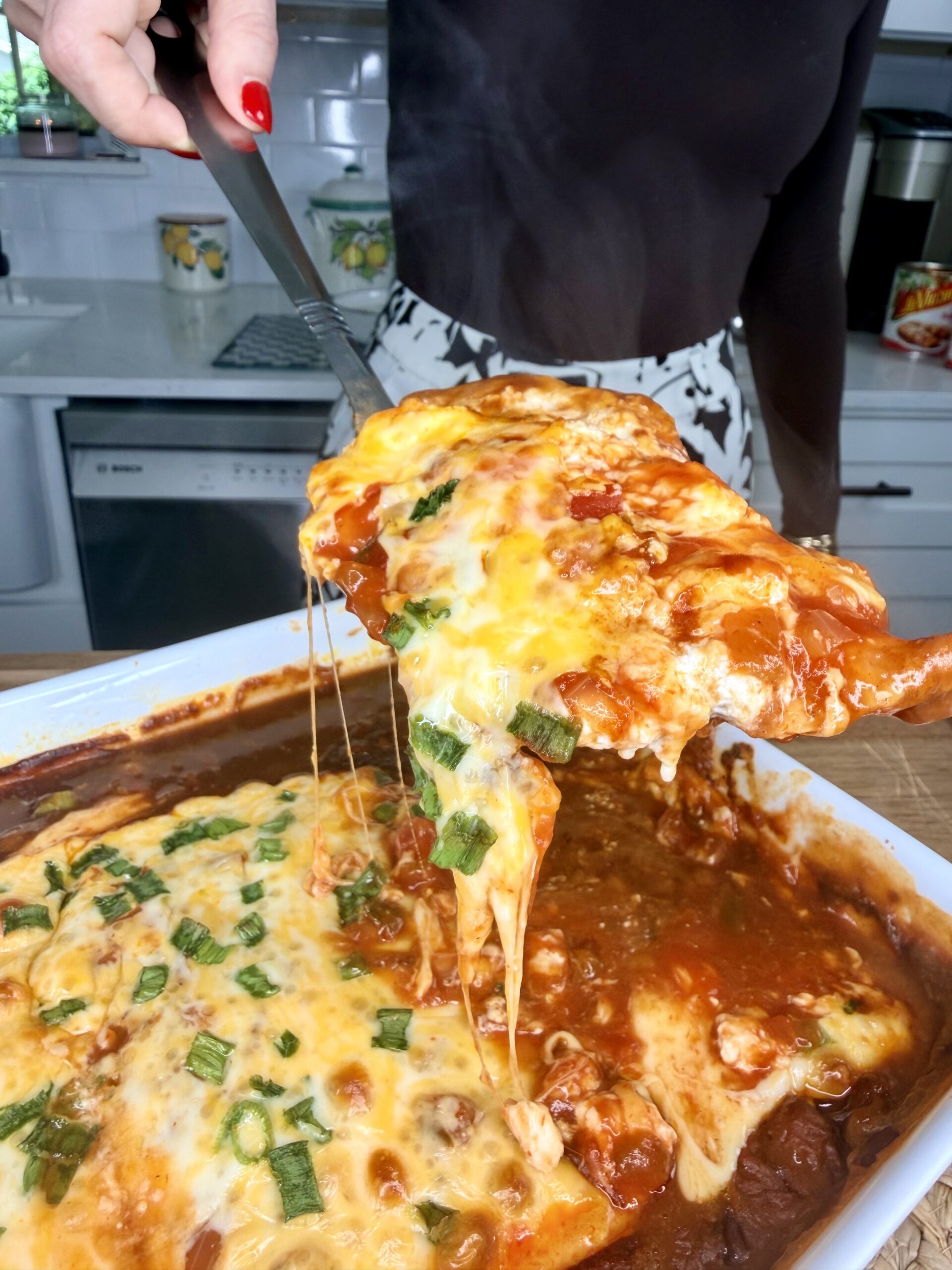 A person serves a cheesy, baked casserole with tomato sauce and green onions from a white dish, with melted cheese stretching from the pan. A kitchen is visible in the background.