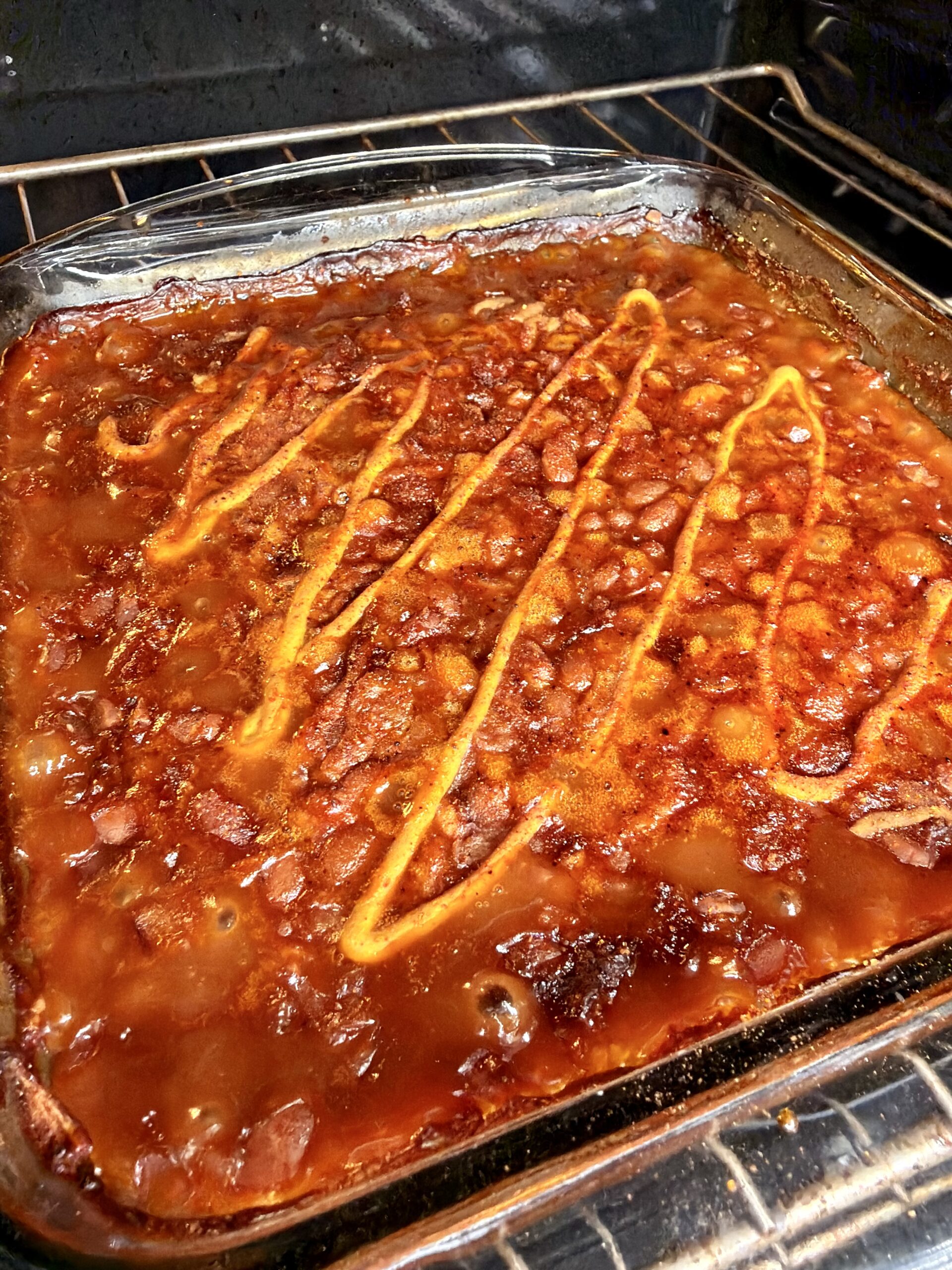 A glass baking dish filled with baked beans in a thick, reddish sauce is shown in an oven, topped with a zigzag drizzle of yellow mustard.