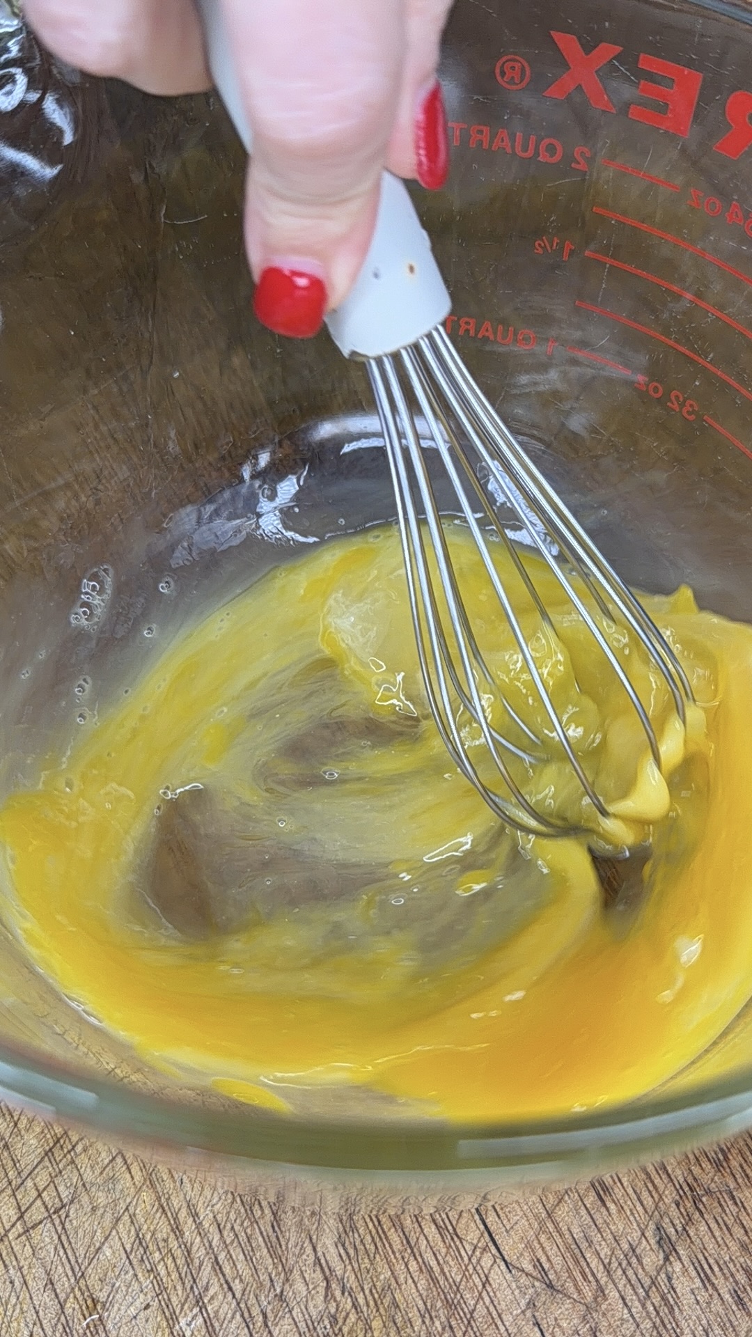 A hand with red nail polish is whisking eggs in a clear glass bowl on a wooden surface.