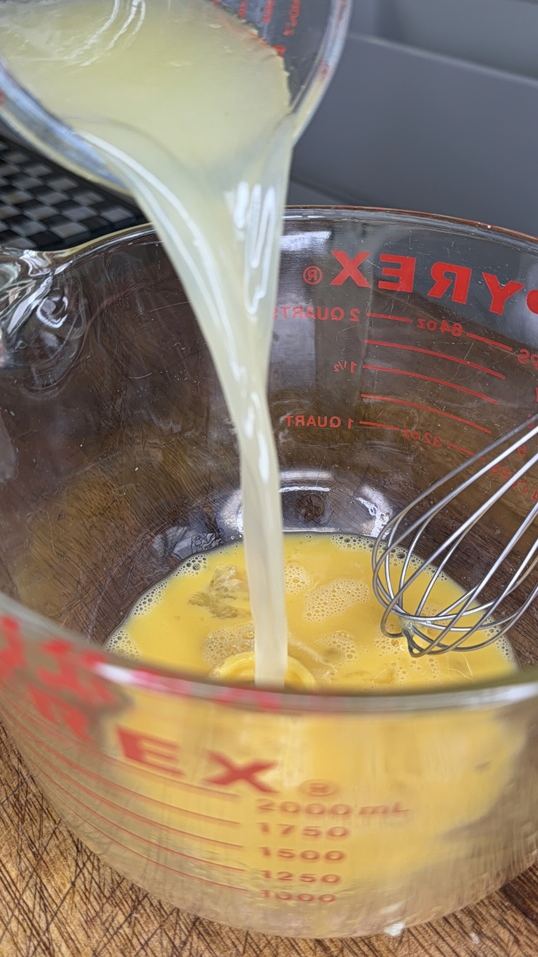 Lemon juice is being poured from a small glass container into a Pyrex measuring cup with beaten eggs, next to a metal whisk on a wooden surface.