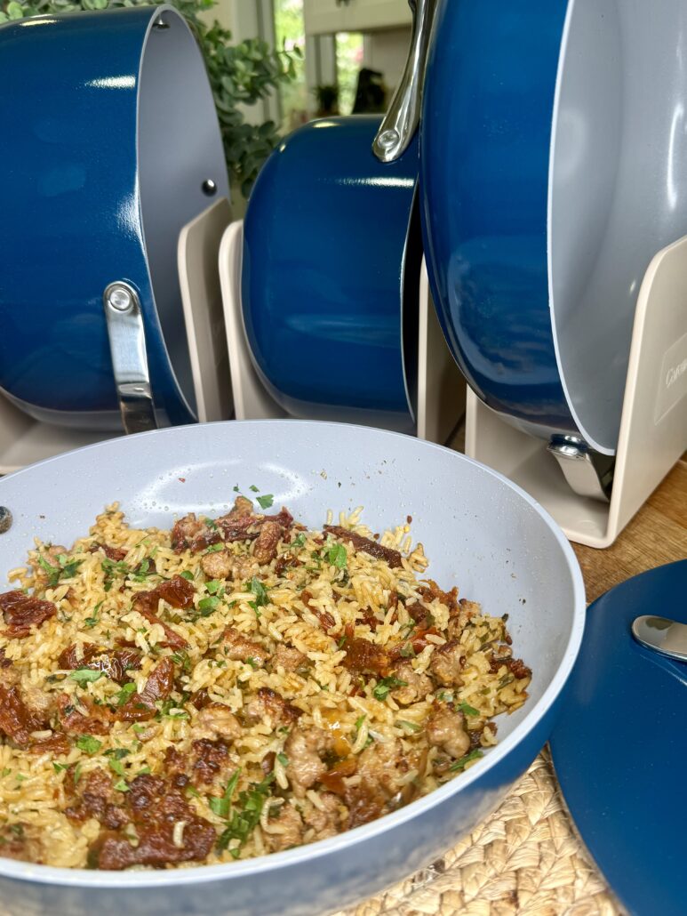 A close-up of a pan filled with cooked rice, ground meat, and herbs sits on a woven placemat, next to a set of blue and gray nonstick cookware arranged upright in a kitchen setting.