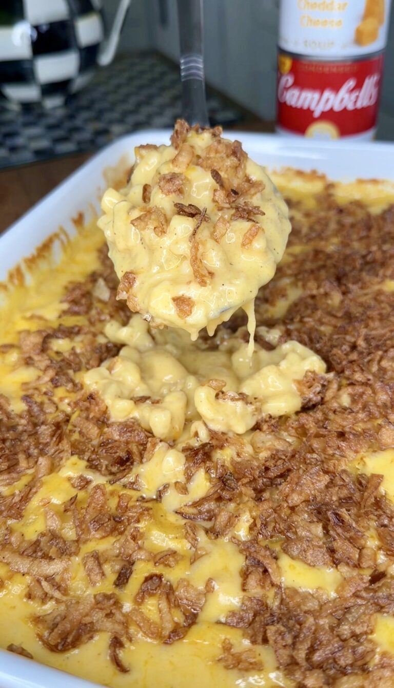 A close-up of a spoonful of creamy macaroni and cheese topped with crispy fried onions, being lifted from a baking dish filled with the same cheesy, golden-brown casserole.
