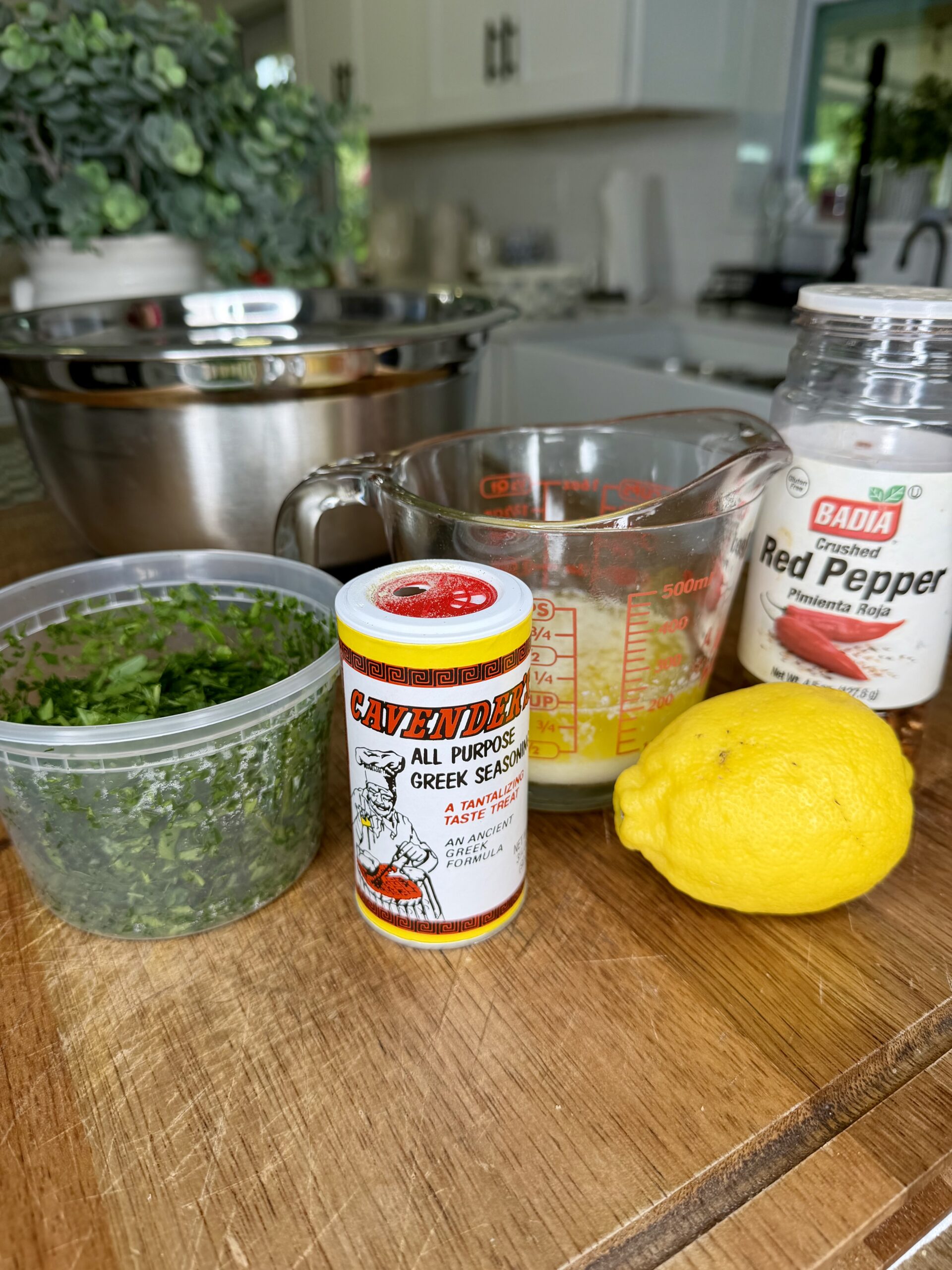 Fresh chopped herbs in a plastic container, a canister of Greek seasoning, a measuring cup with liquid, a lemon, and a jar of crushed red pepper sit on a wooden counter in a bright kitchen.