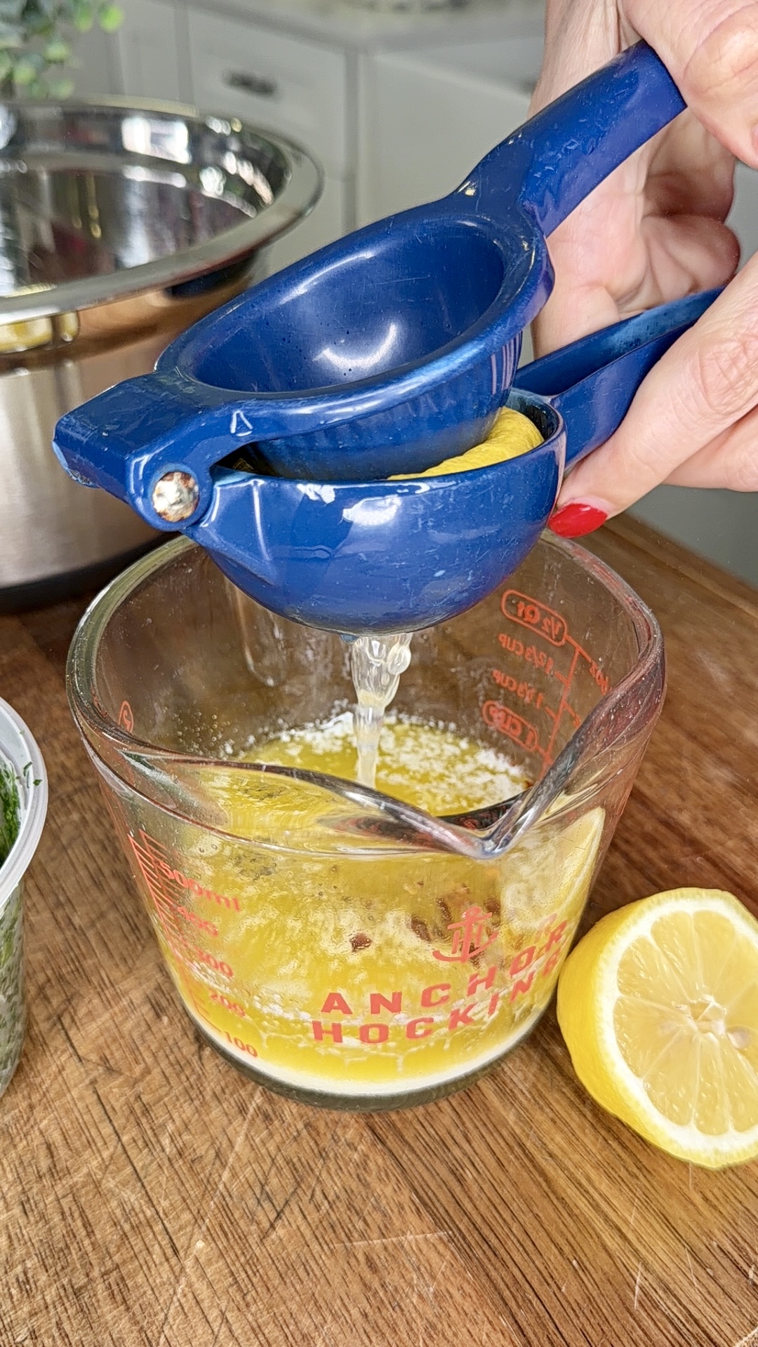 A hand uses a blue citrus squeezer to juice a lemon half over a glass measuring cup filled with liquid. Another lemon half sits nearby on a wooden countertop.