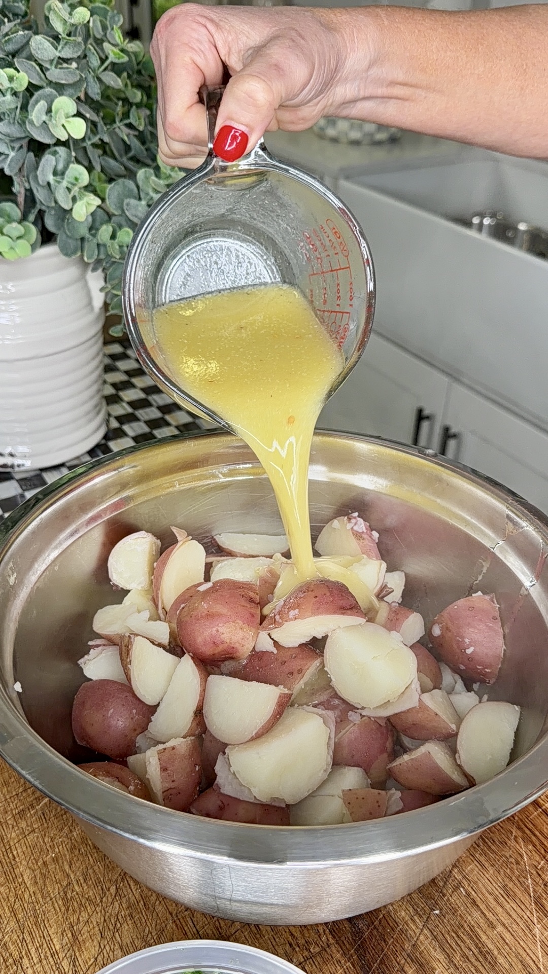 A hand pours melted butter from a glass measuring cup over a bowl of cooked, chopped red potatoes on a wooden counter in a kitchen.