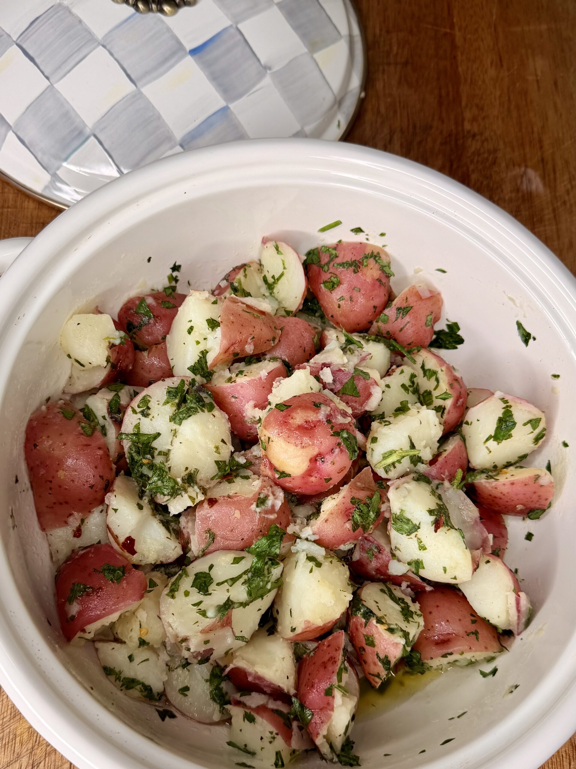 A white ceramic bowl filled with cooked red potatoes cut into chunks, tossed with chopped parsley and herbs, on a wooden surface next to a checkered lid.