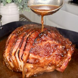 A ladle pours brown glaze over a roasted ham with crispy, caramelized edges, sitting in a pan of sauce. Steam rises from the meat, and a plant and kitchen sink are visible in the blurred background.