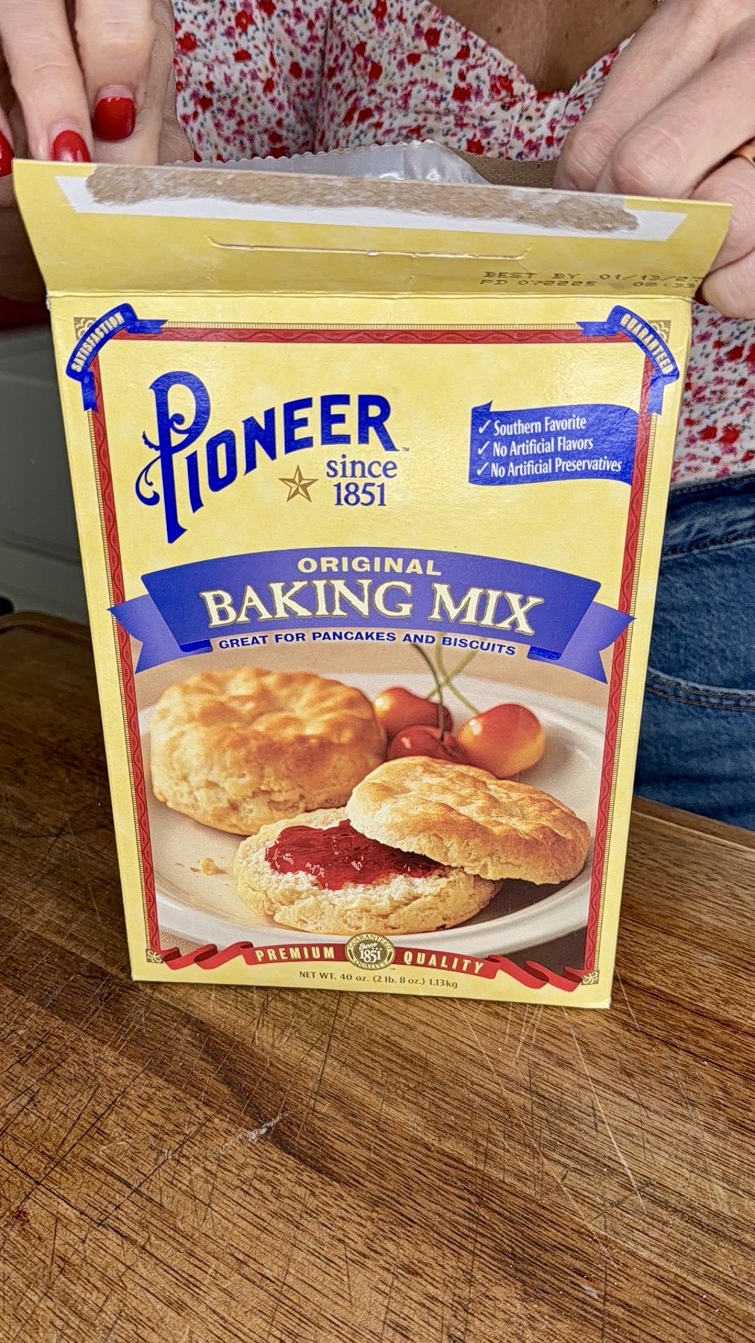 A person holds an open box of Pioneer Original Baking Mix, which is labeled as great for pancakes and biscuits, on a wooden surface. The box shows images of biscuits with jam and the brands logo.