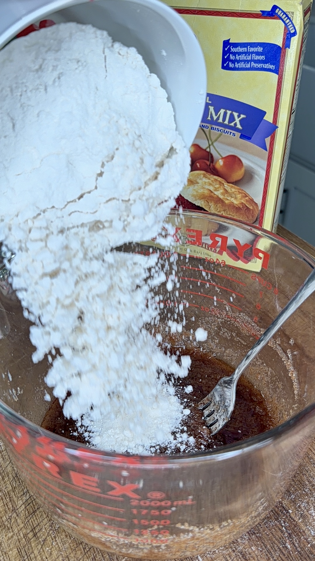 A bowl of flour is being poured into a glass mixing bowl containing a brown liquid mixture, with a fork inside. A box of baking mix is visible in the background.