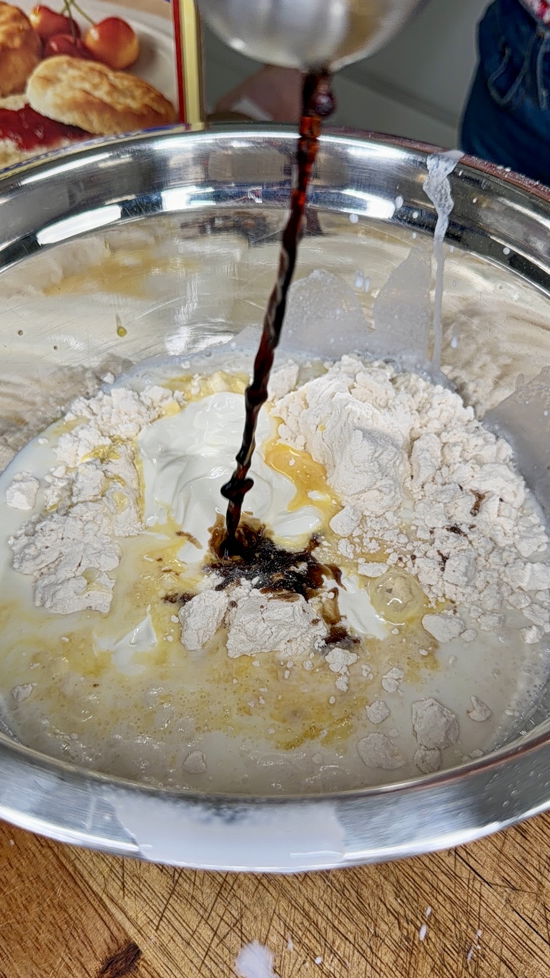 A close-up of a metal bowl with flour, sour cream, and other baking ingredients being mixed, while a dark liquid is poured in, likely for making biscuits. A biscuit mix box is visible in the background.