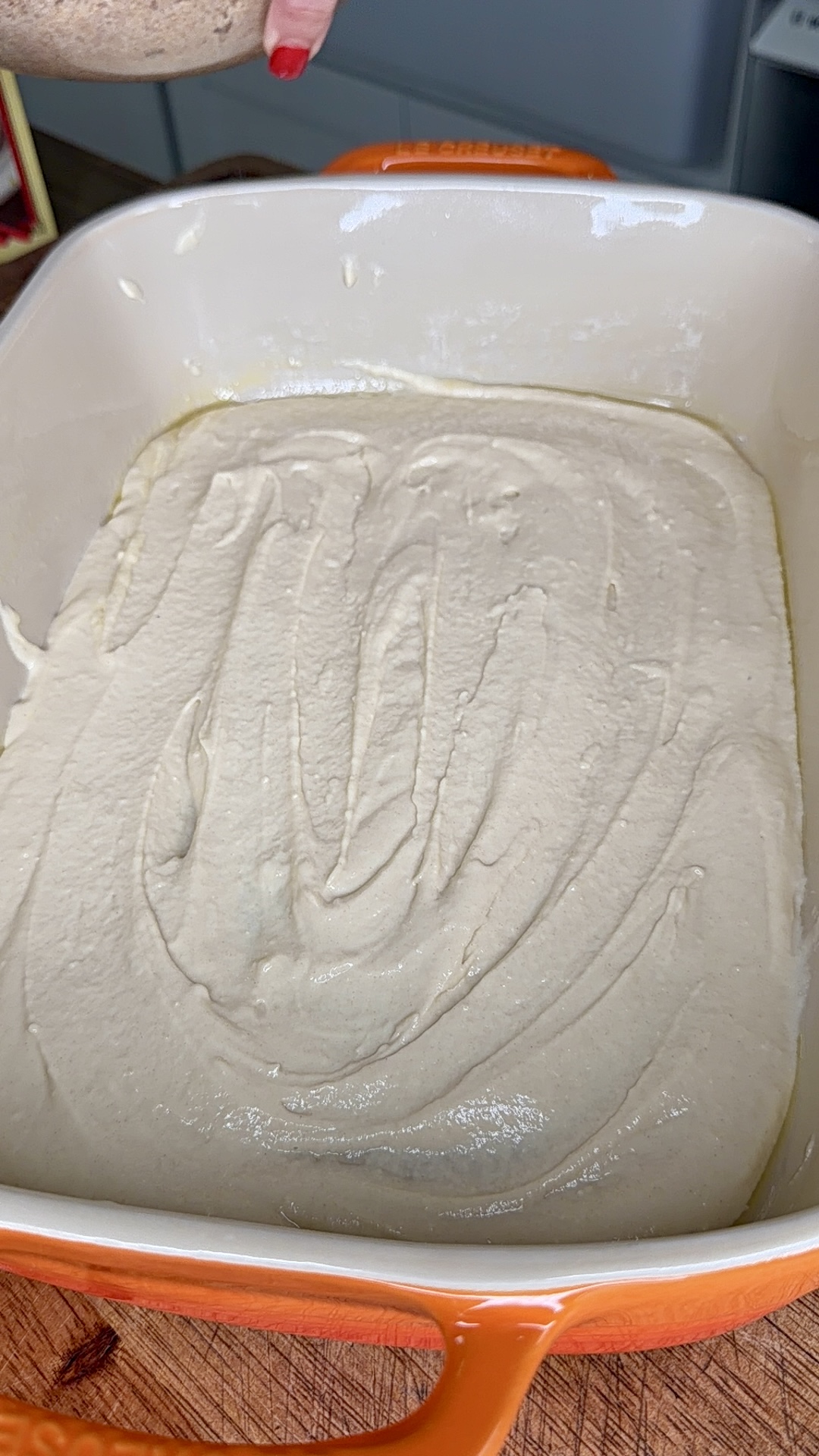 A baking dish with a creamy, light batter spread evenly inside, ready for baking. The dish is orange and sits on a wooden surface. A hand with red nail polish is visible at the top.