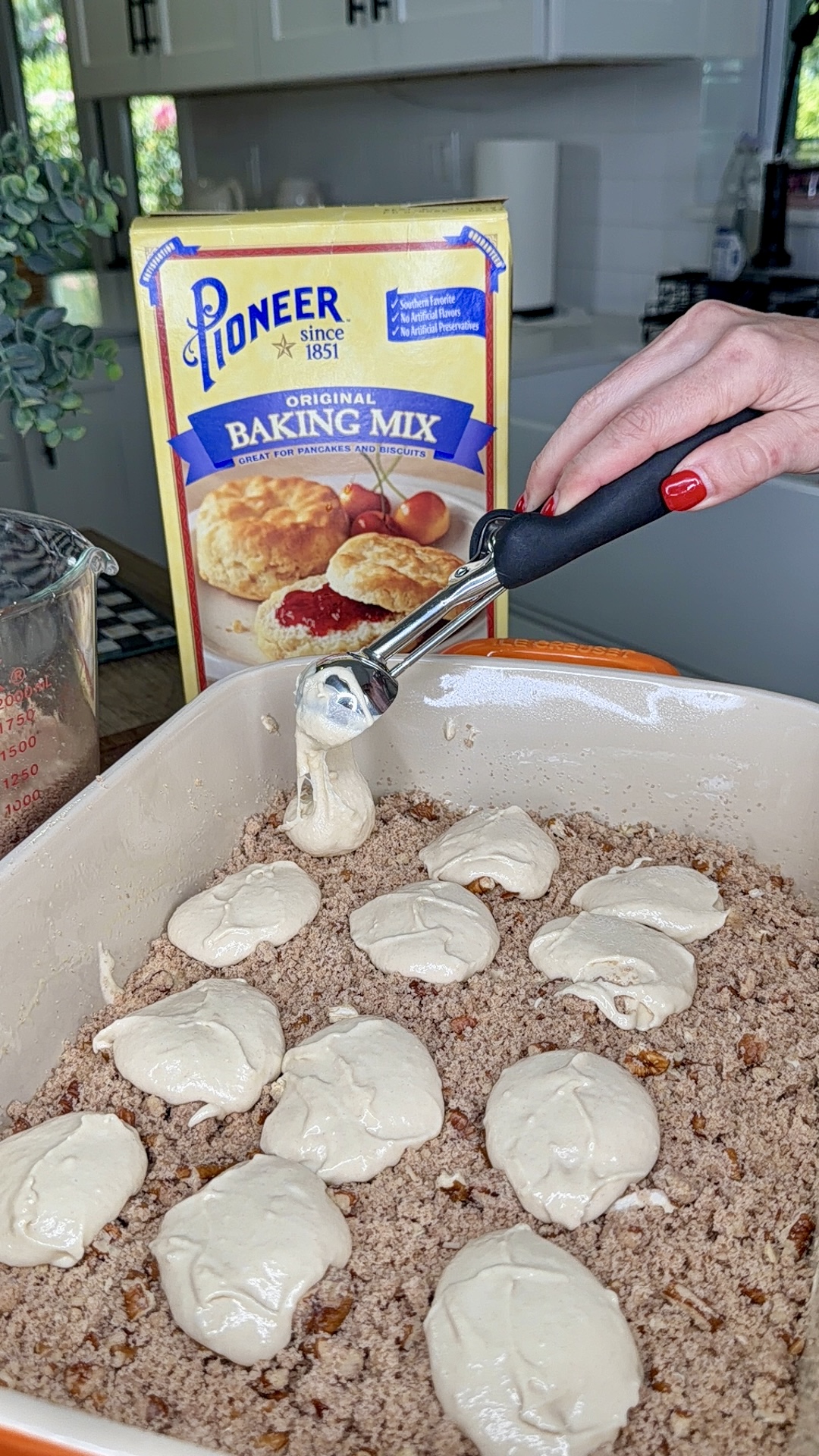 A hand uses a scoop to place biscuit dough over a crumbly mixture in a baking dish. Behind the dish is a box of Pioneer Original Baking Mix on a kitchen counter.