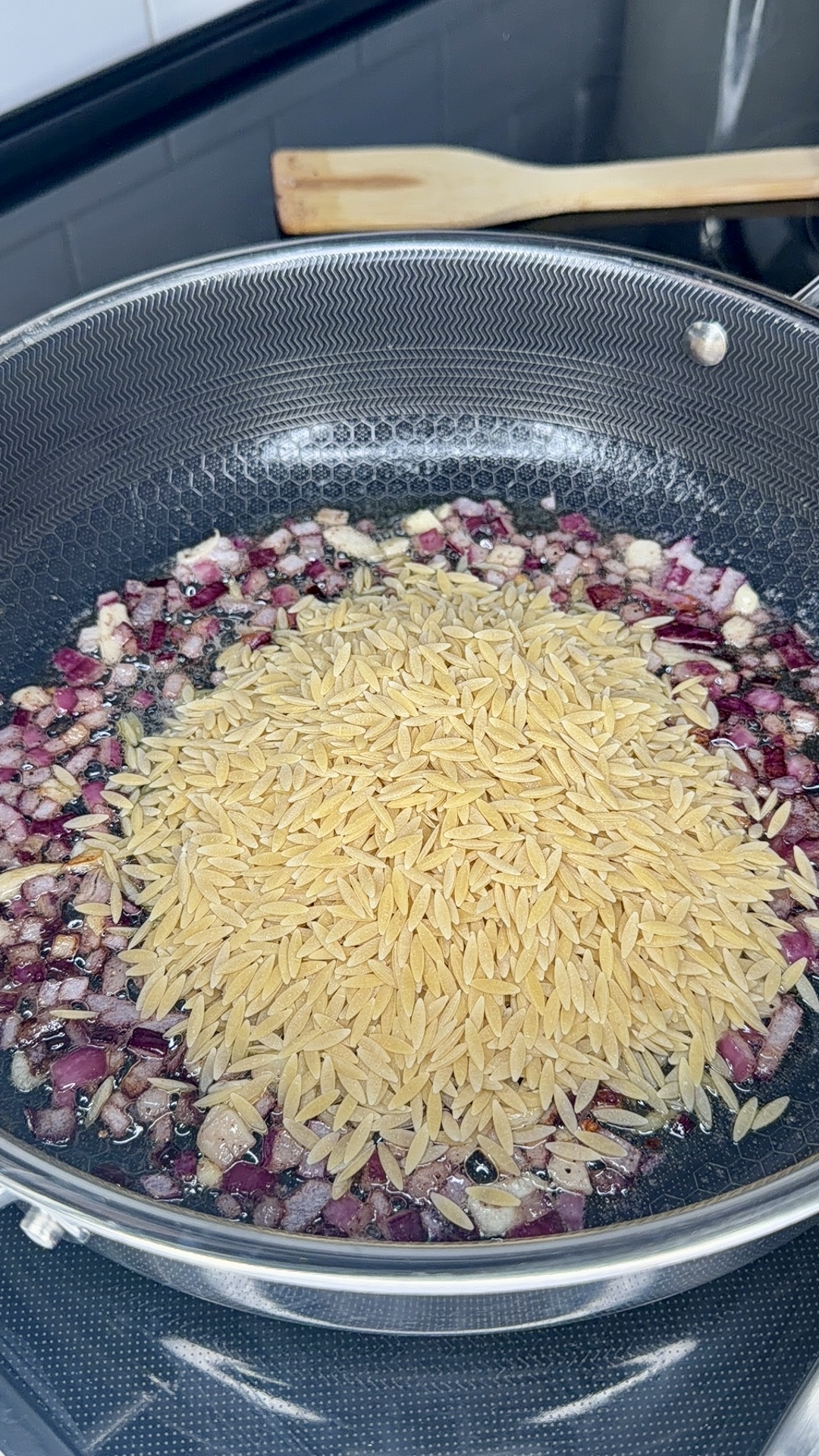 A frying pan on a stovetop contains chopped red onions being sautéed in oil, with uncooked orzo pasta piled on top. A wooden spatula rests on the counter in the background.