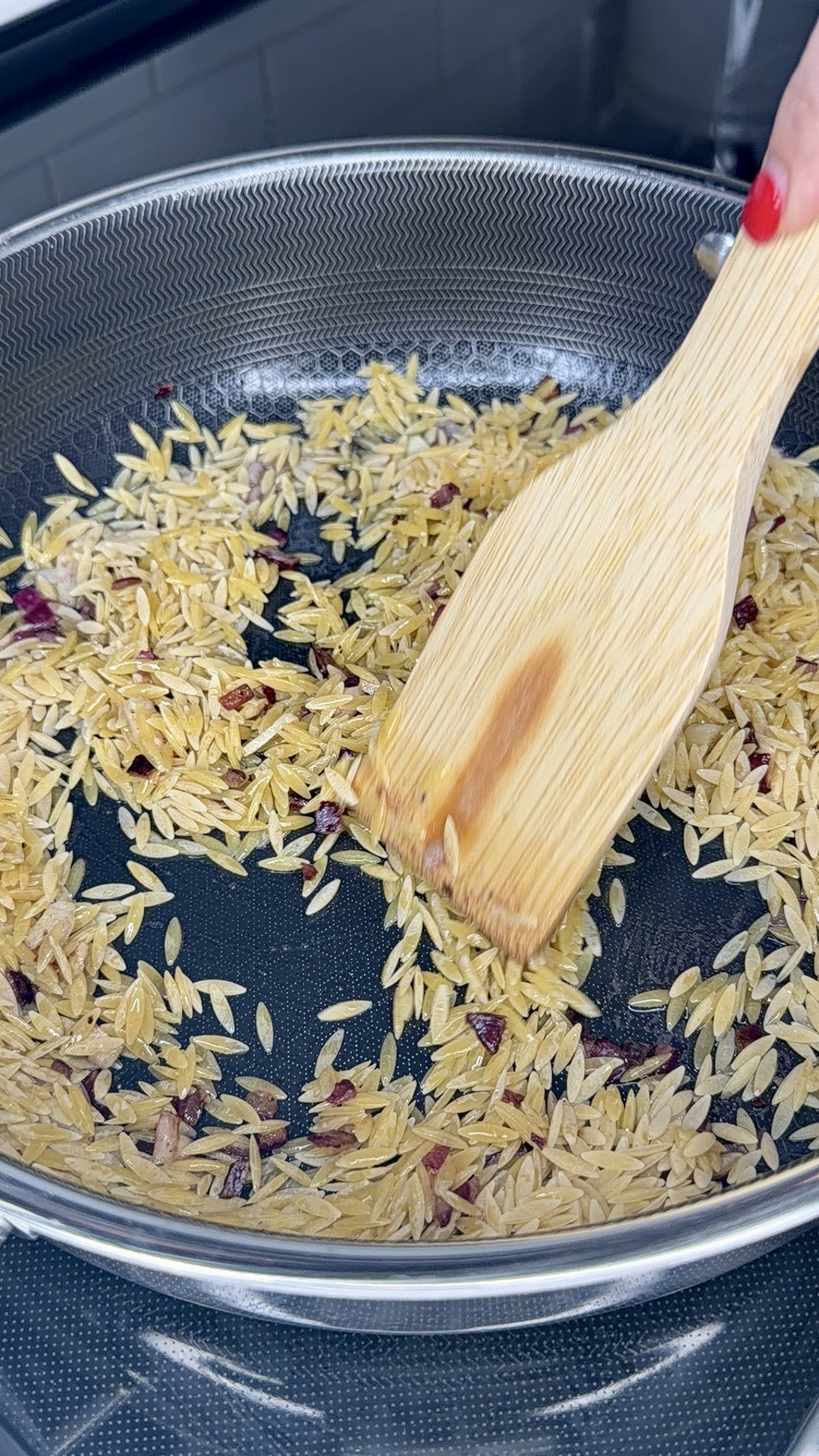 A hand with red nail polish stirs One-Pan Sicilian Orzo and diced onions in a frying pan using a wooden spatula. The orzo is being toasted and is starting to turn golden brown.