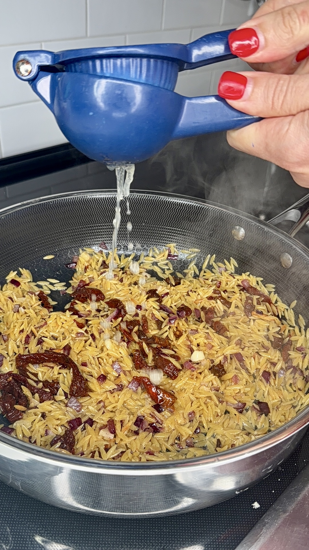 A hand with red nail polish squeezes a blue citrus juicer over a pan of orzo pasta mixed with sun-dried tomatoes and onions on a stovetop. Steam is rising from the pan.