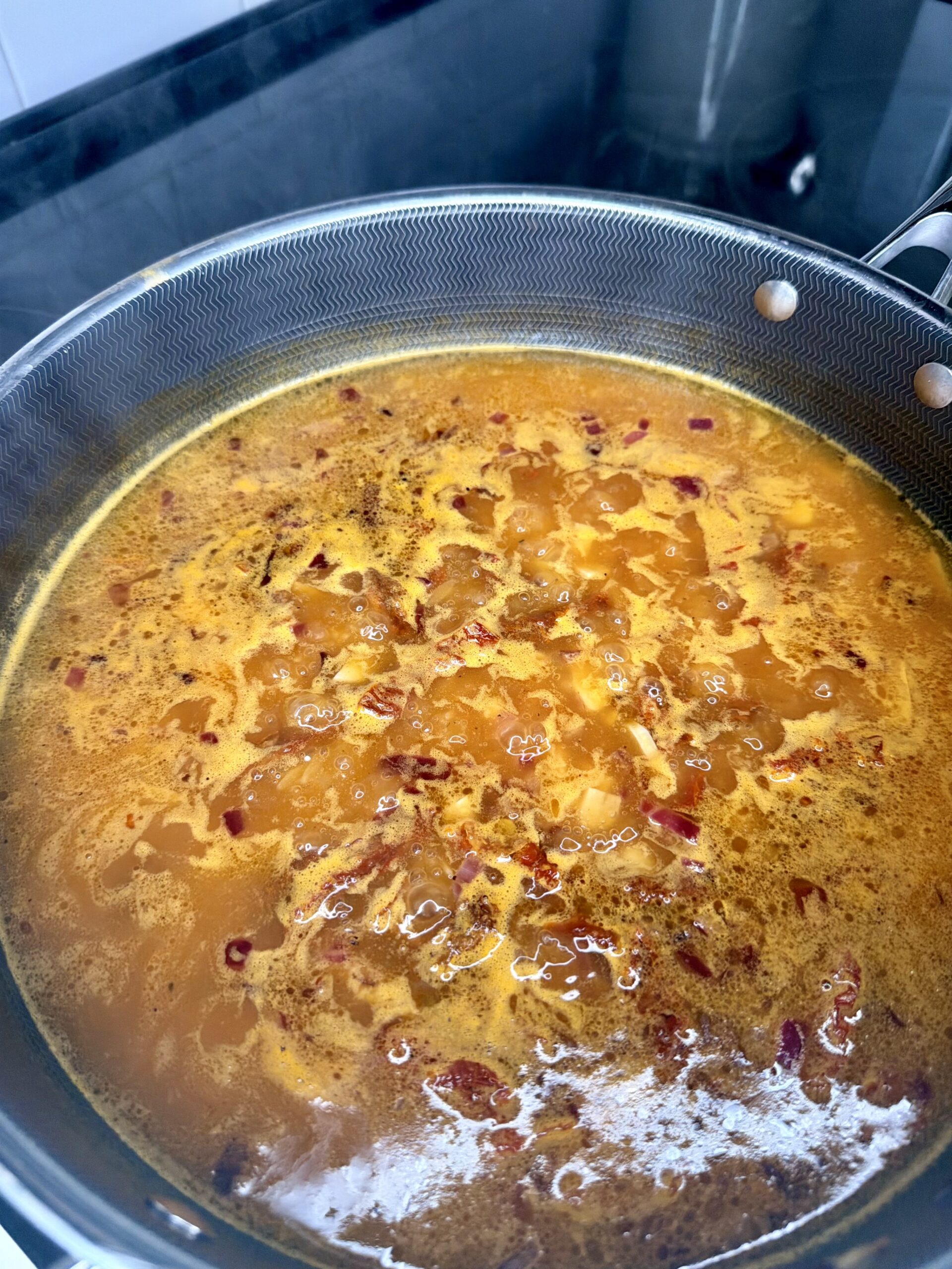 A close-up of a pot on a stove filled with simmering orange-red One-Pan Sicilian Orzo, featuring chopped onions and red peppers. The bubbling liquid suggests the flavorful dish is being cooked or boiled.