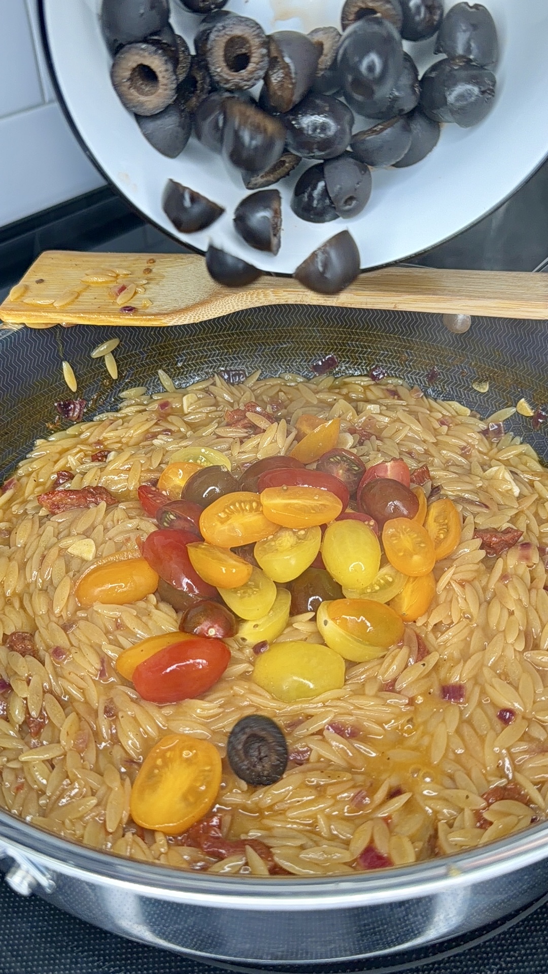 A bowl of sliced black olives is being poured into a pan of cooked One-Pan Sicilian Orzo mixed with halved cherry tomatoes in various colors. Steam rises from the pan, and a wooden spatula rests on the edge, ready to stir everything together.