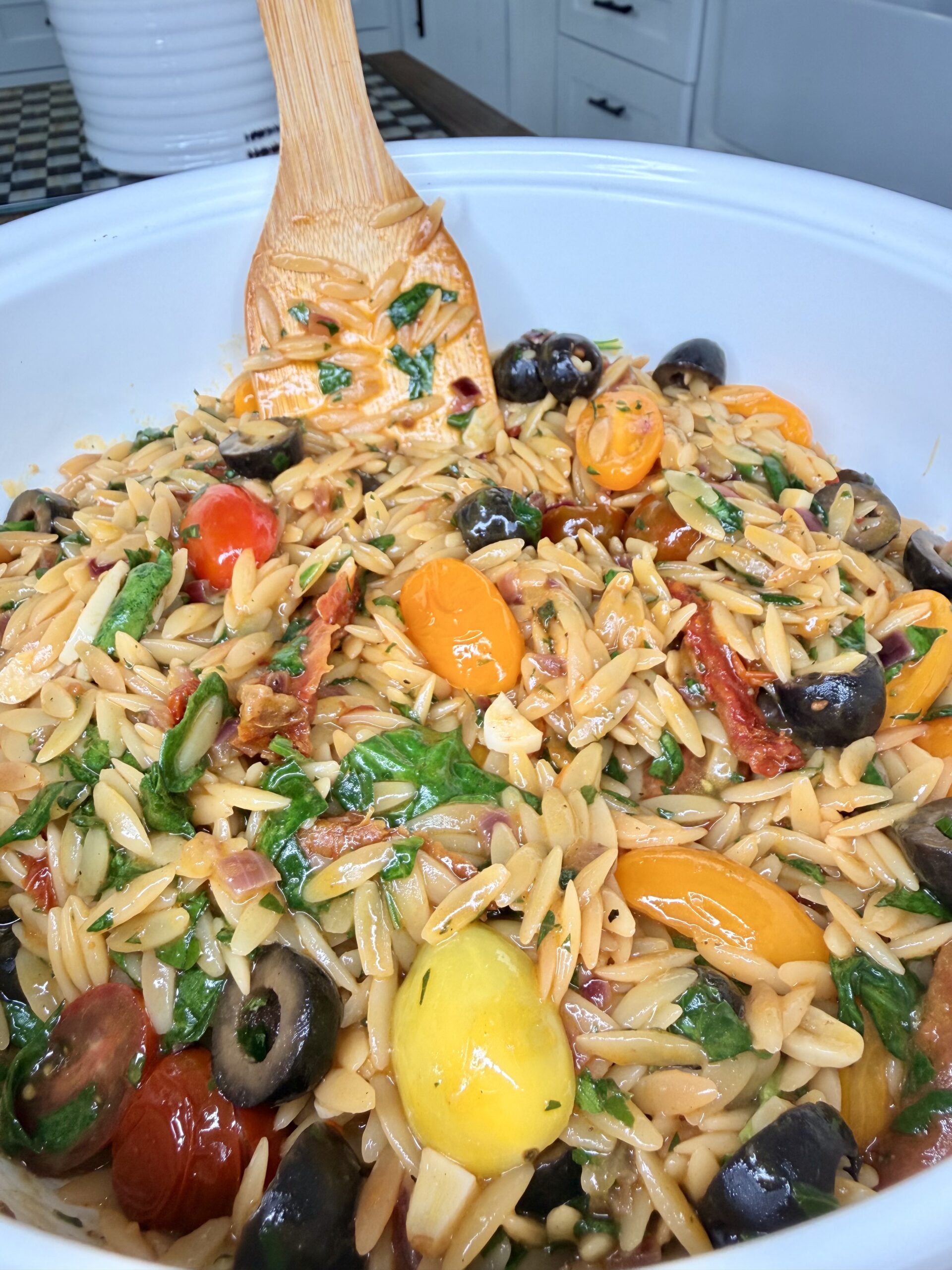 A close-up of a white bowl filled with orzo pasta salad, featuring cherry tomatoes, black olives, spinach, sun-dried tomatoes, and a wooden spoon for mixing.