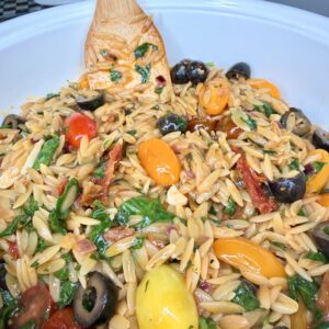 A close-up of a large bowl filled with orzo pasta salad mixed with cherry tomatoes, black olives, spinach, sun-dried tomatoes, and a wooden spoon for stirring.