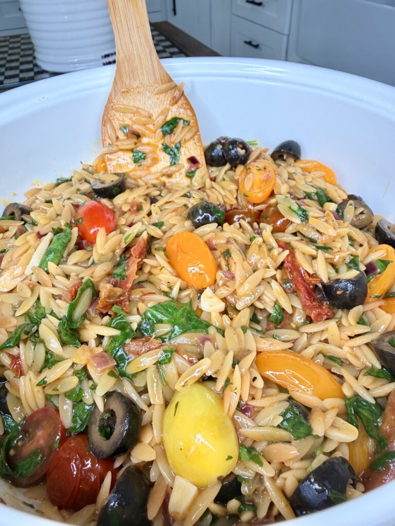 A close-up of a large bowl filled with orzo pasta salad mixed with cherry tomatoes, black olives, spinach, sun-dried tomatoes, and a wooden spoon for stirring.