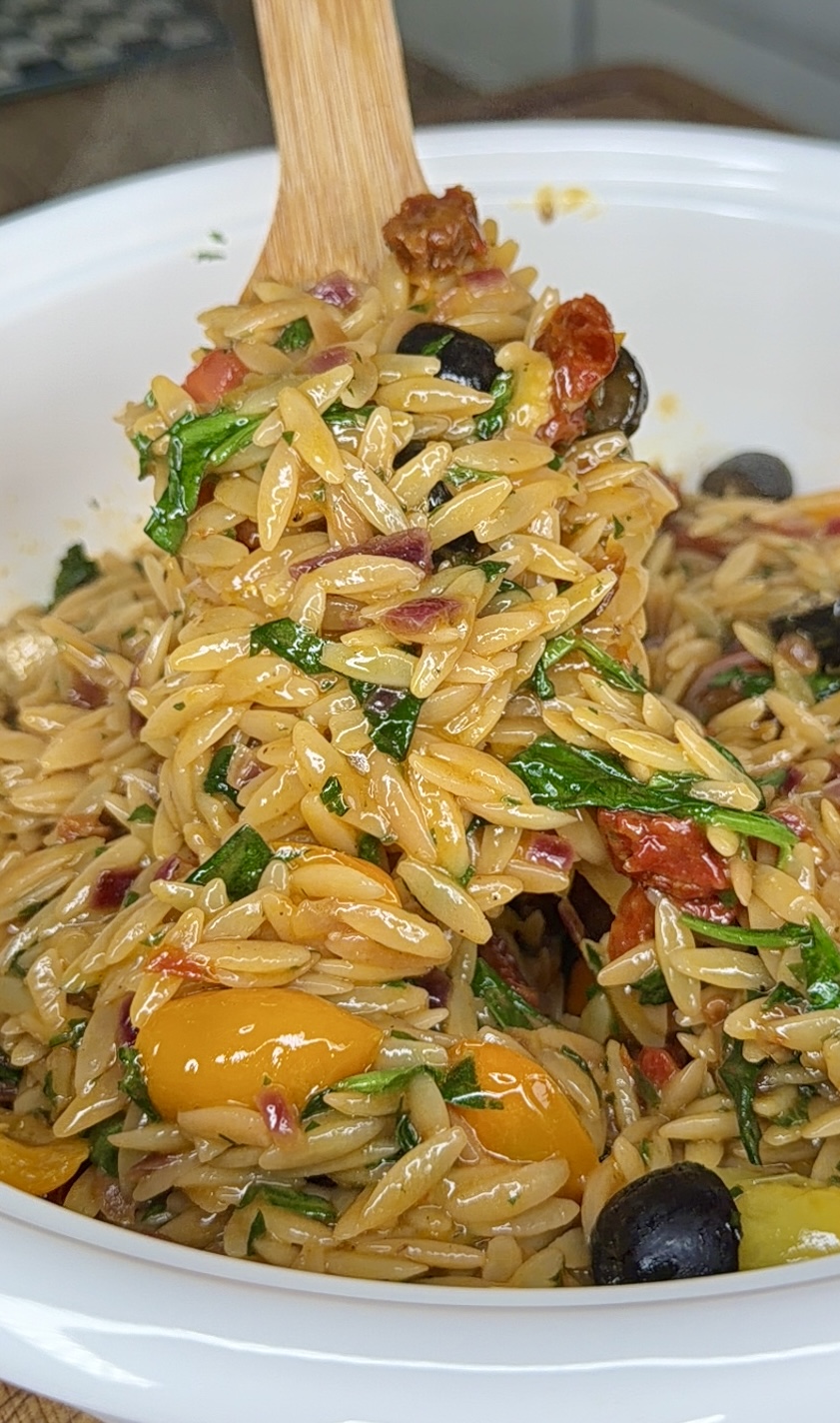 A close-up of a spoonful of orzo pasta salad with spinach, black olives, cherry tomatoes, sun-dried tomatoes, red onion, and herbs in a white bowl.