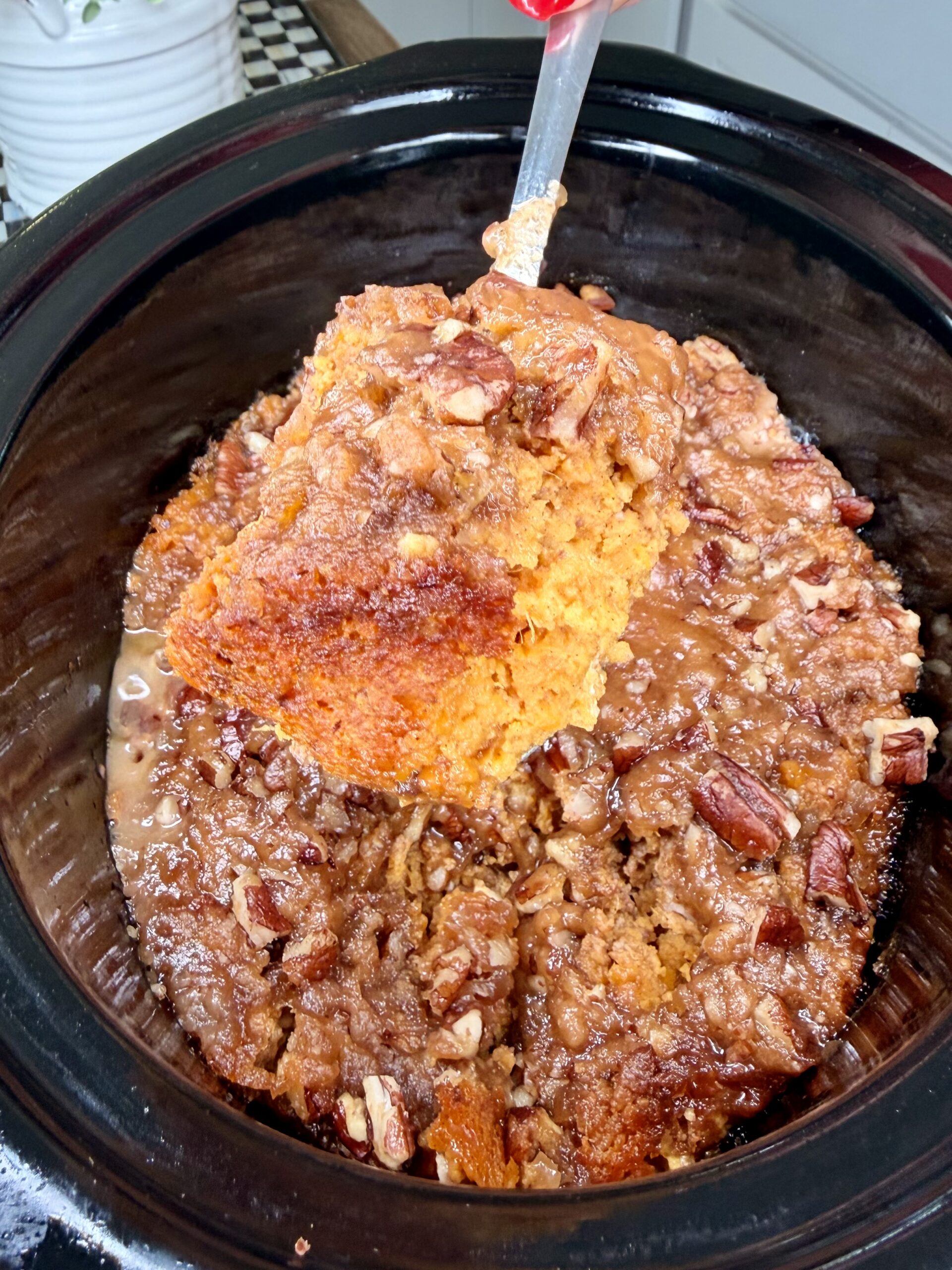 A piece of pumpkin pecan cobbler is being lifted out of a slow cooker with a spatula, its moist, orange interior and crumbly pecan topping resembling the cozy appeal of a Crockpot Sweet Potato Casserole.