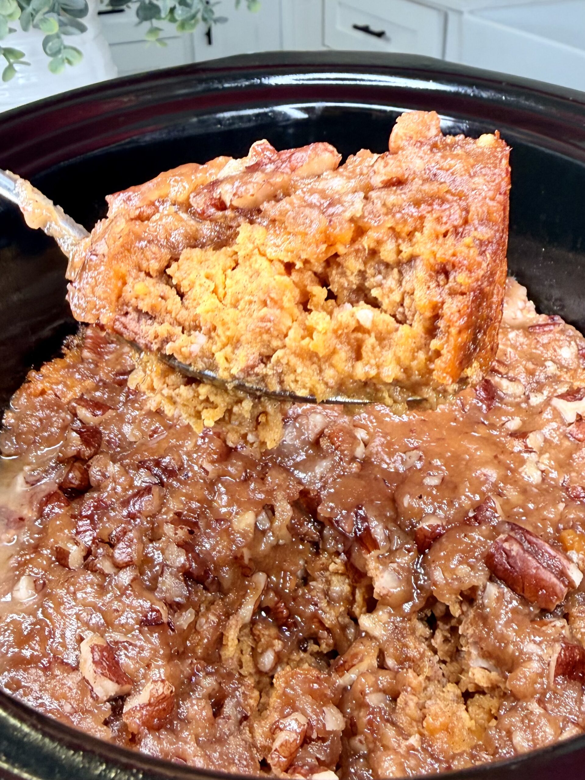 A close-up of a spoon lifting a serving of moist Crockpot Sweet Potato Casserole from a slow cooker, revealing its crumbly, golden-orange interior and nutty, caramelized pecan topping.