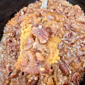 A close-up of Crockpot Sweet Potato Casserole with a golden-brown top, chopped pecans, and a moist texture, being lifted from a baking dish with a metal spatula.