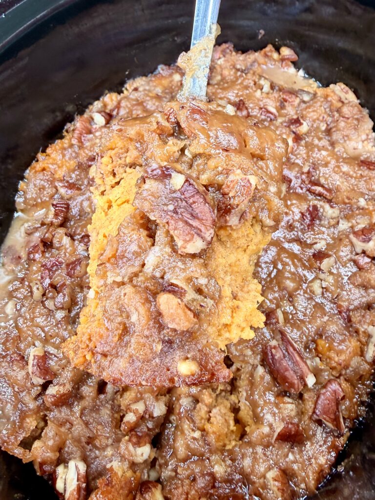 A close-up of Crockpot Sweet Potato Casserole with a golden-brown top, chopped pecans, and a moist texture, being lifted from a baking dish with a metal spatula.