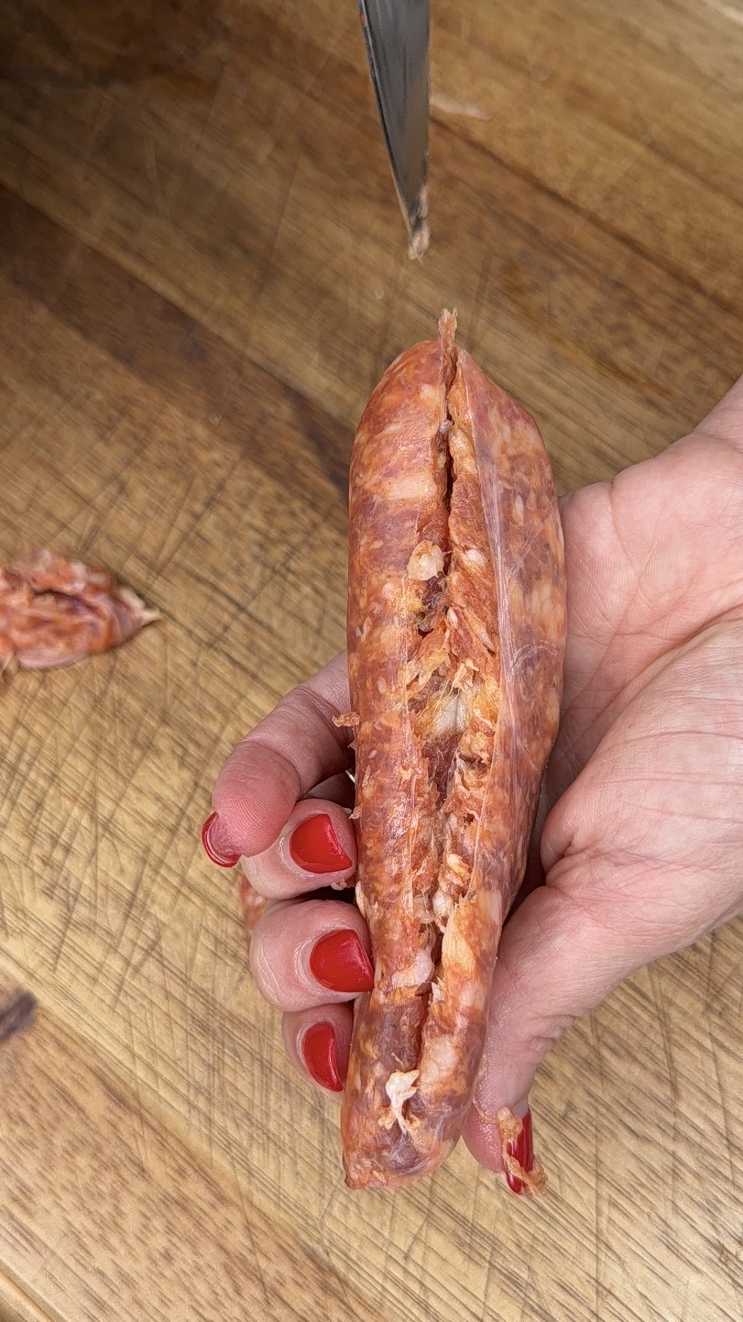 A hand with red-painted nails holds a partially sliced cured sausage on a wooden cutting board. A knife is visible at the top, cutting into the sausage.