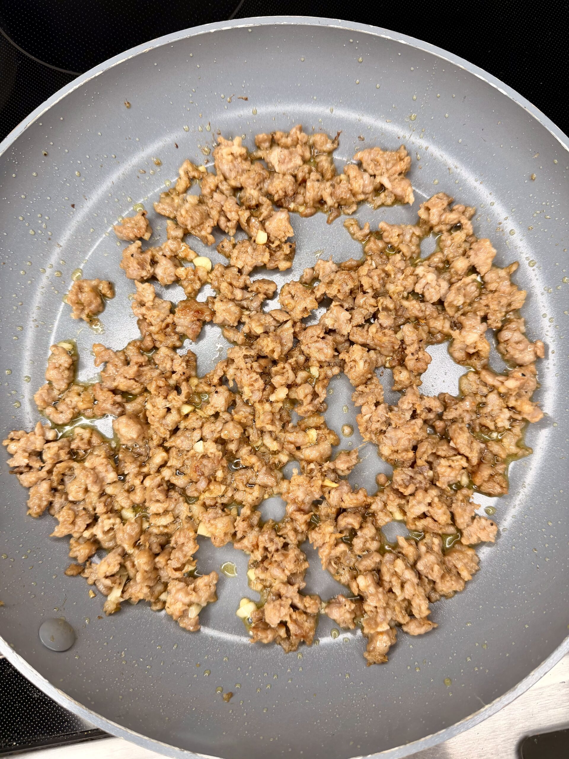 Ground meat is being browned and cooked with small bits of garlic and seasoning in a nonstick frying pan on a stovetop. The meat is spread out evenly across the pan.