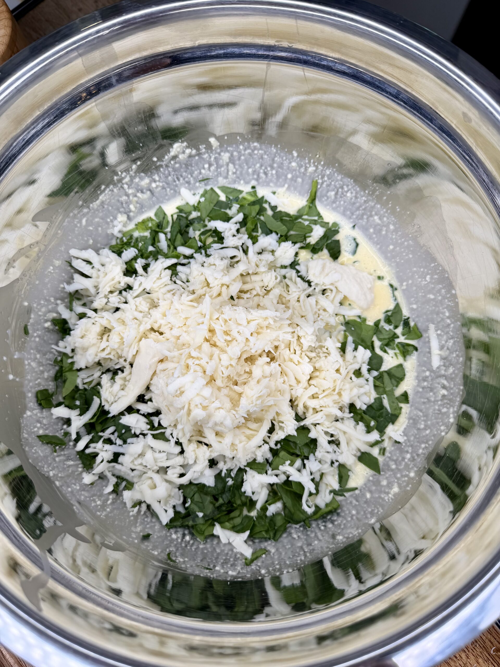 A metal mixing bowl containing shredded cheese, chopped spinach, and liquid ingredients, ready to be mixed together.