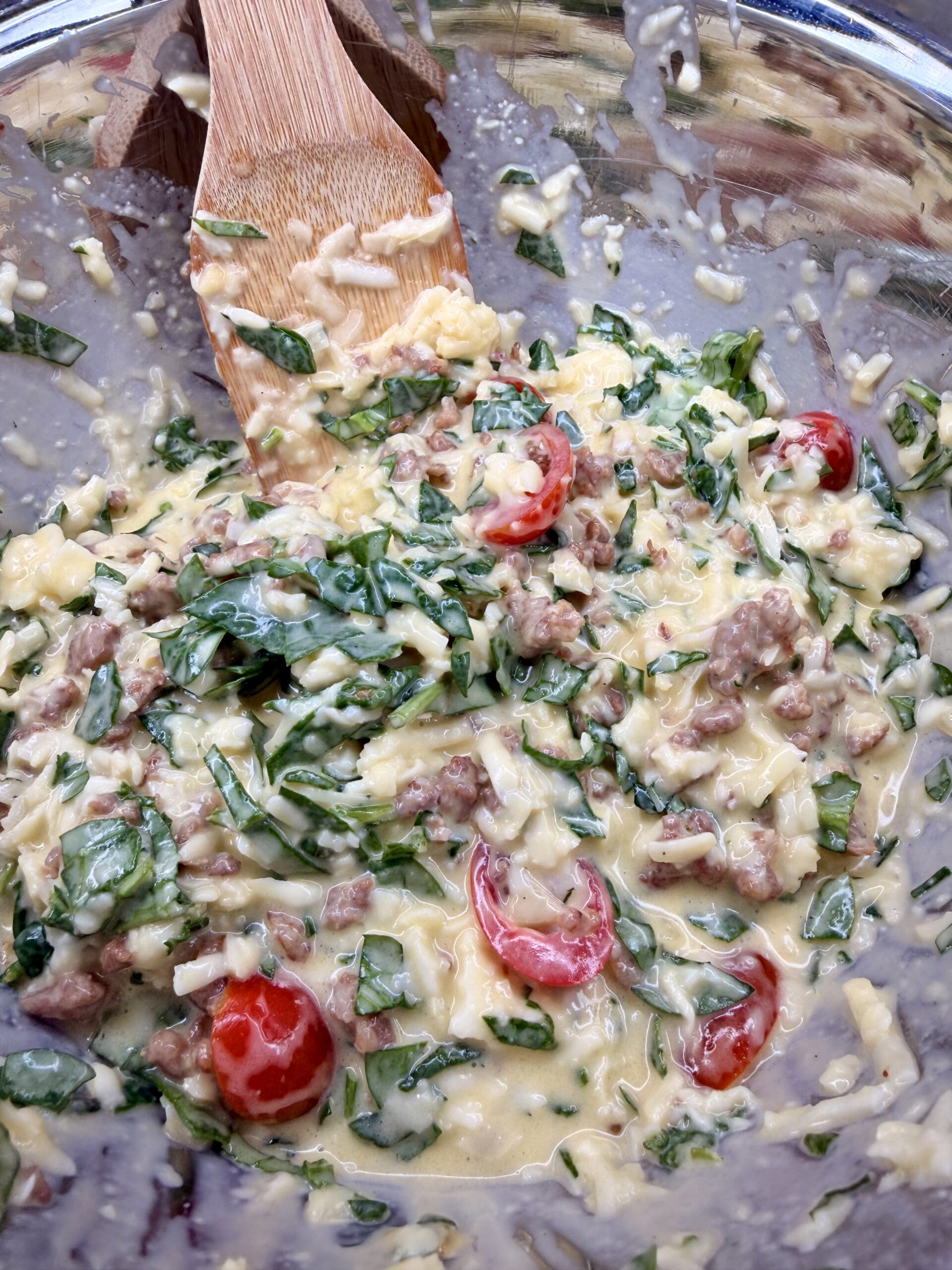 A close-up of a mixing bowl filled with a creamy mixture of chopped spinach, cherry tomatoes, ground sausage, and shredded cheese being stirred with a wooden spatula.