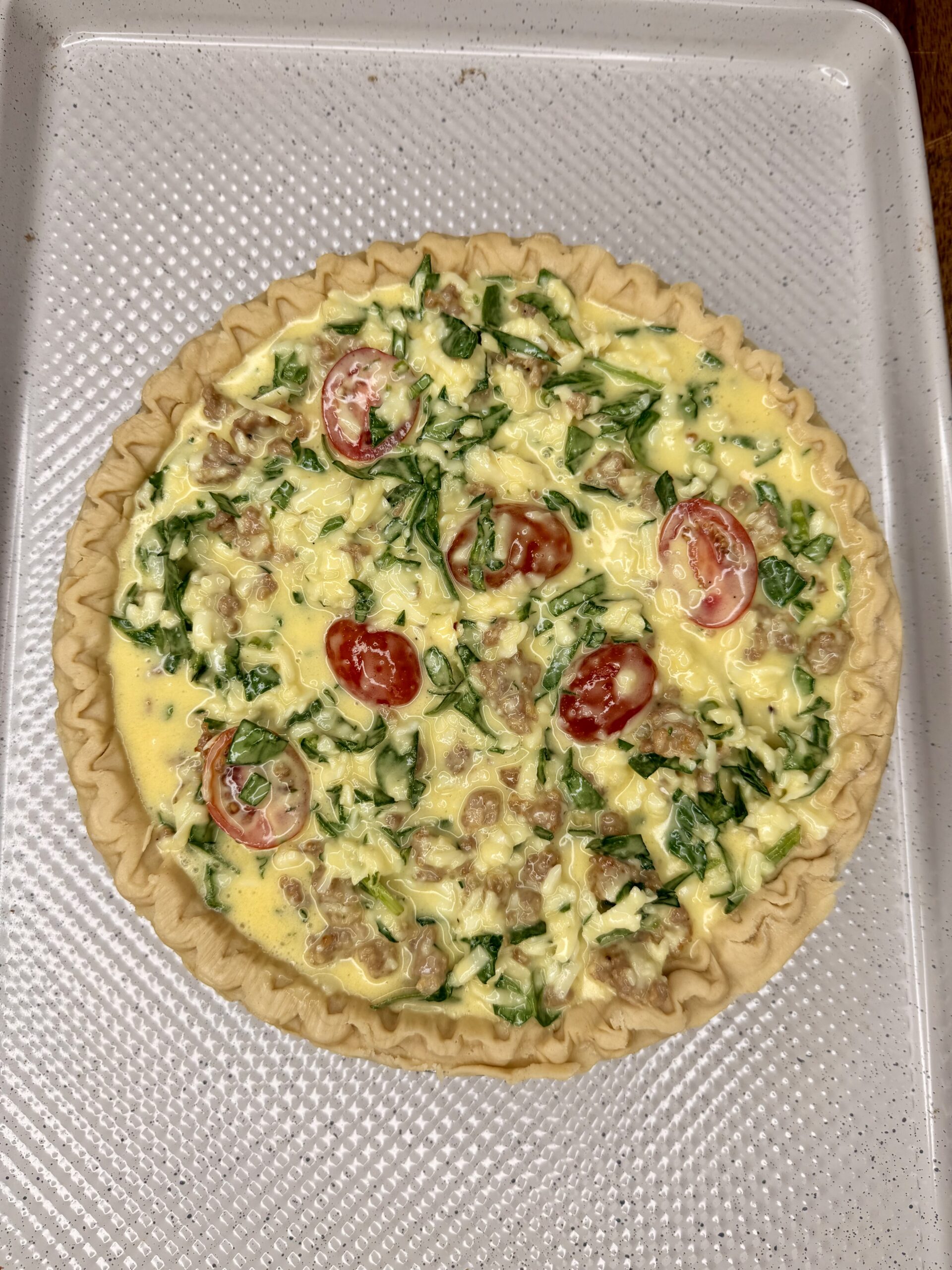 Unbaked quiche in a pie crust on a baking tray, filled with eggs, spinach, sausage, shredded cheese, and halved cherry tomatoes.