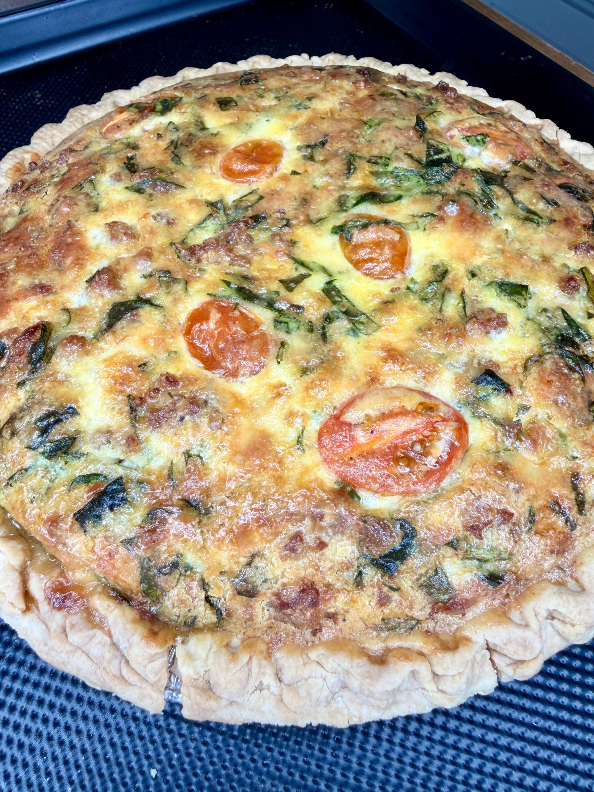 A freshly baked quiche with a golden-brown crust, topped with slices of tomato and visible pieces of green vegetables, resting on a dark textured surface.