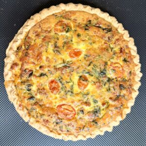 A whole baked quiche with a golden crust, filled with visible spinach, melted cheese, and halved cherry tomatoes, photographed from above on a black textured background.