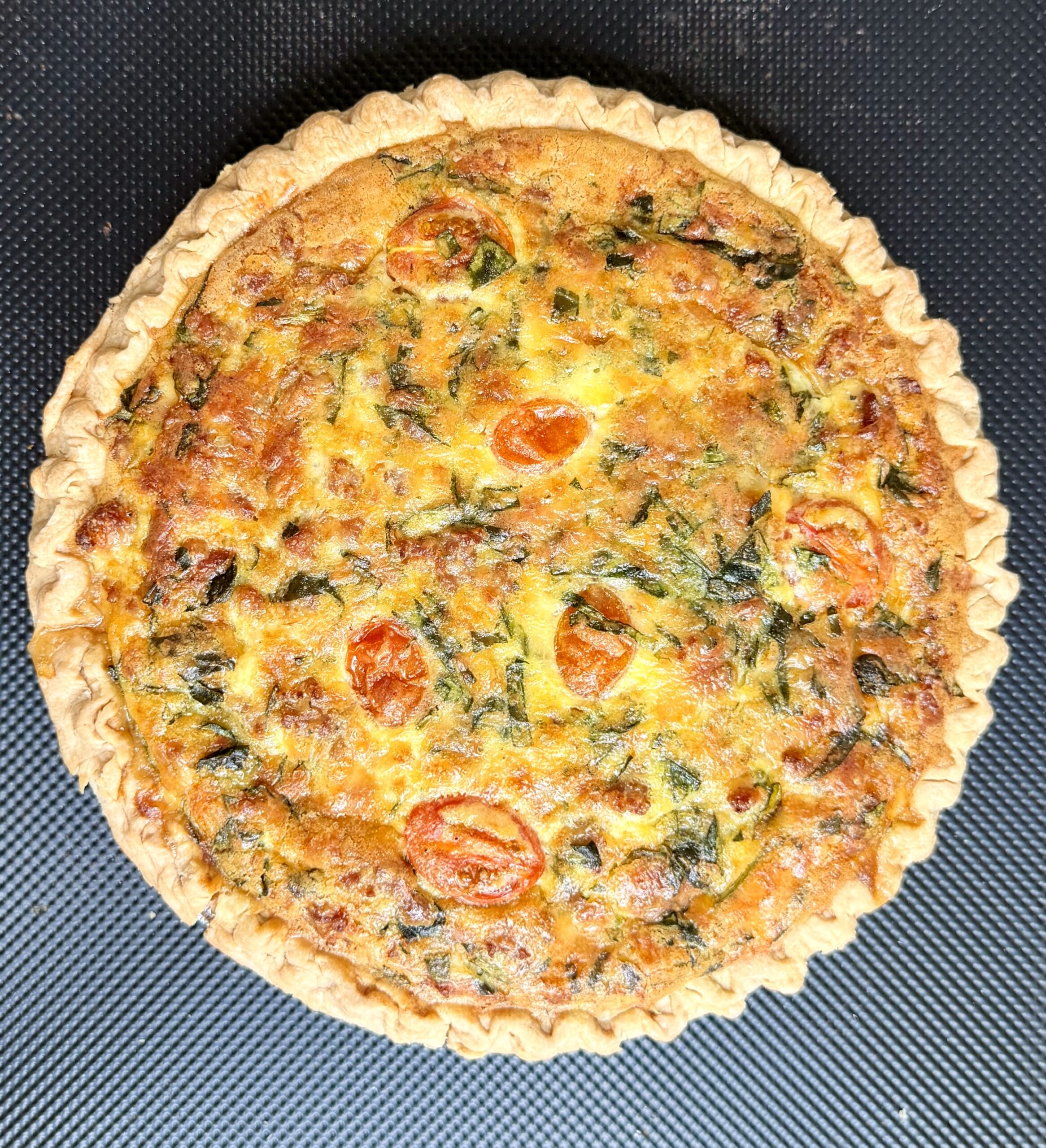 A whole baked quiche with a golden crust, filled with visible spinach, melted cheese, and halved cherry tomatoes, photographed from above on a black textured background.