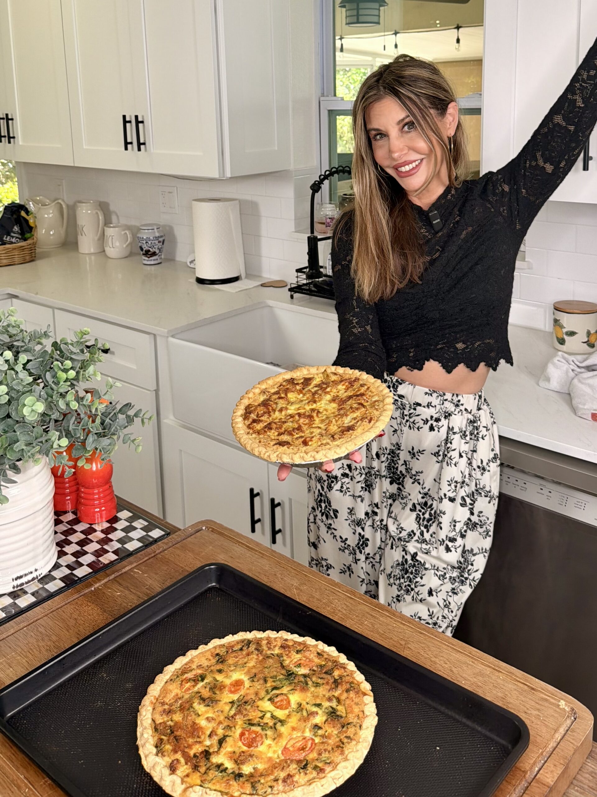 A smiling woman in a black lace top and patterned pants holds a freshly baked quiche in a kitchen. Another quiche sits on a baking tray in front of her. White cabinets and a farmhouse sink are in the background.