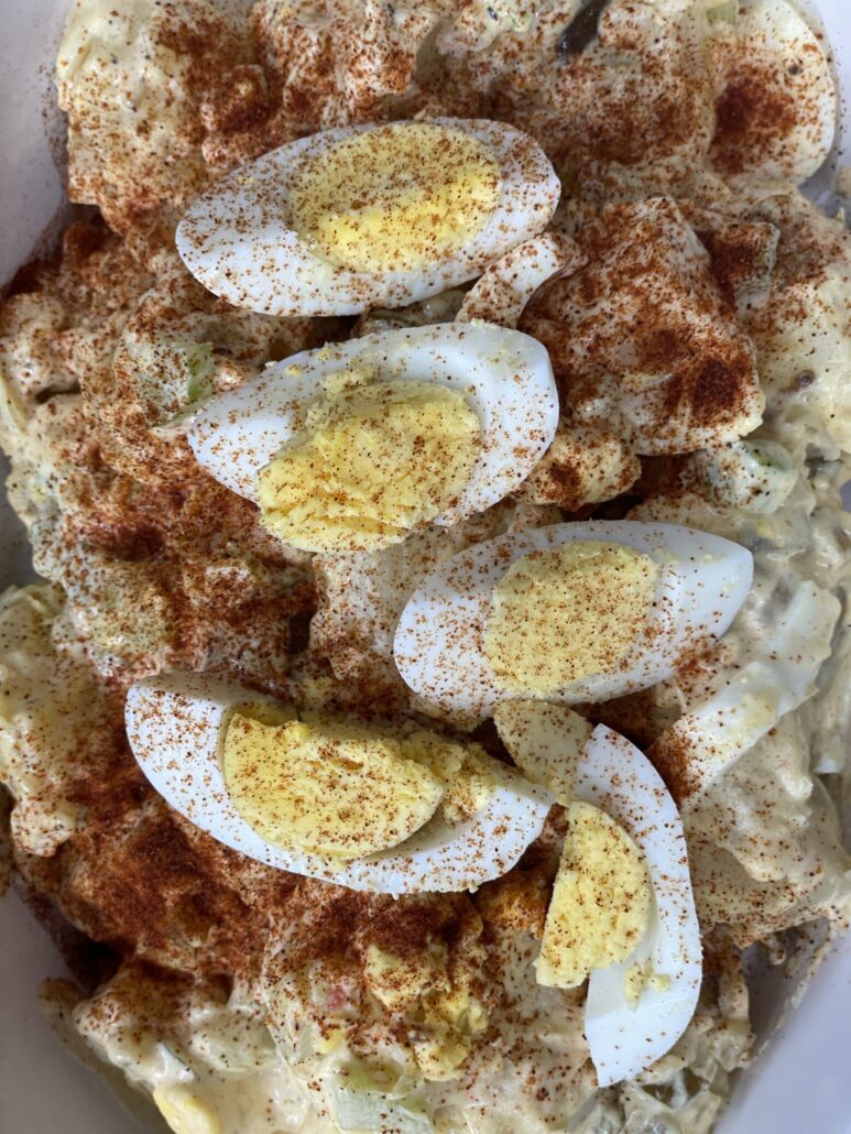 Close-up of creamy potato salad topped with sliced hard-boiled eggs and a generous sprinkle of paprika, served in a white dish.