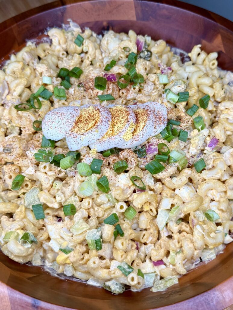 Close-up of a bowl of macaroni salad topped with slices of hard-boiled egg, sprinkled with paprika, and garnished with chopped green onions. The salad contains diced celery and red onions.
