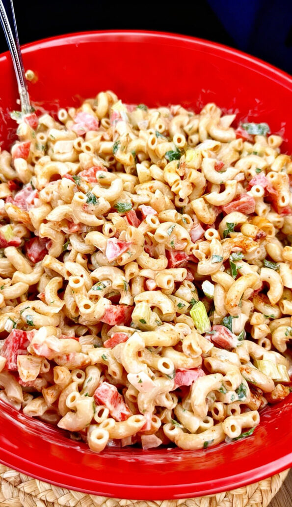 A close-up of a red bowl filled with creamy macaroni salad, featuring elbow pasta, diced red bell peppers, green herbs, and a creamy dressing, with a metal spoon partially visible.