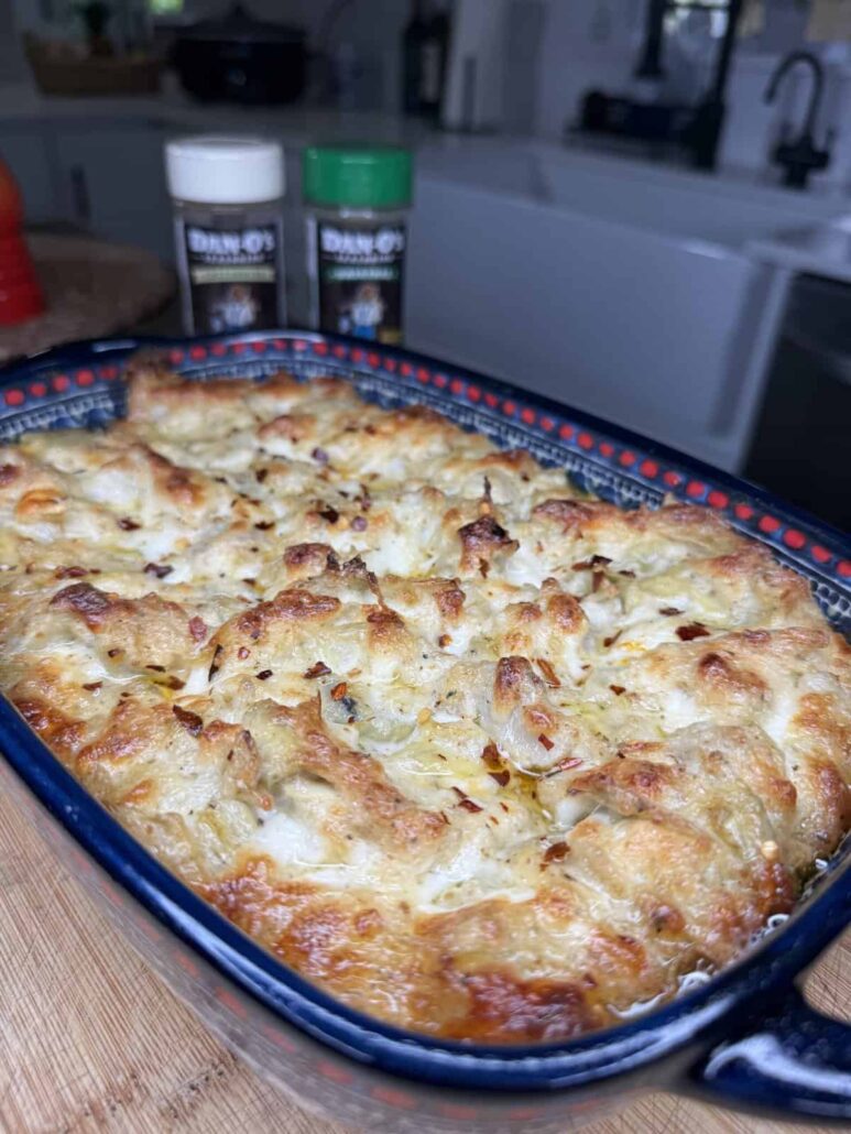 A freshly baked cheesy casserole sits in a blue patterned dish on a wooden surface, with a golden-brown, bubbly topping. Two seasoning containers are visible in the background on a kitchen counter.