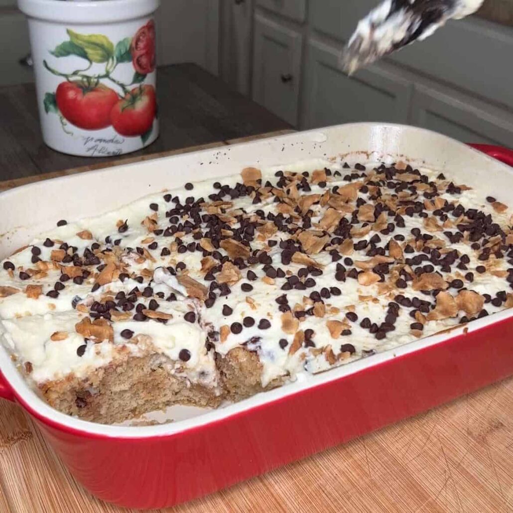 A baking dish with a frosted cake topped with chocolate chips and chopped nuts, one slice removed. A hand holds a serving utensil with cake—perfect for The Ultimate Easter Menu (Mediterranean & American). Tomato-art ceramic container in the background.