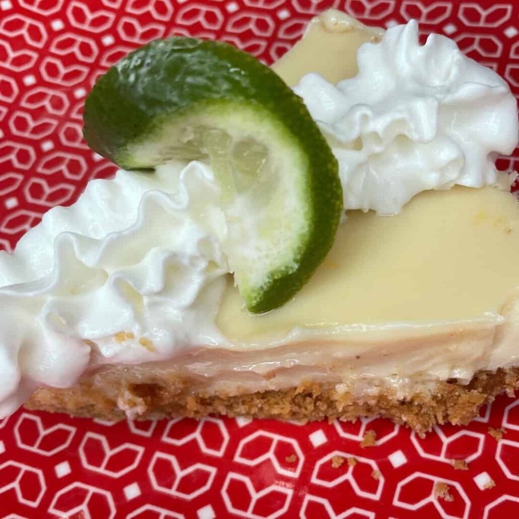 A slice of key lime pie with a graham cracker crust, topped with whipped cream and a lime wedge, on a red plate—perfect for The Ultimate Easter Menu (Mediterranean & American).