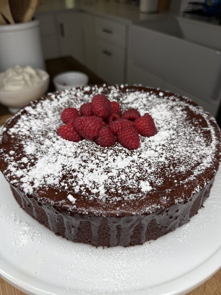 A chocolate cake topped with fresh raspberries and dusted with powdered sugar sits on a white plate, perfect for The Ultimate Easter Menu (Mediterranean & American), in a modern kitchen.
