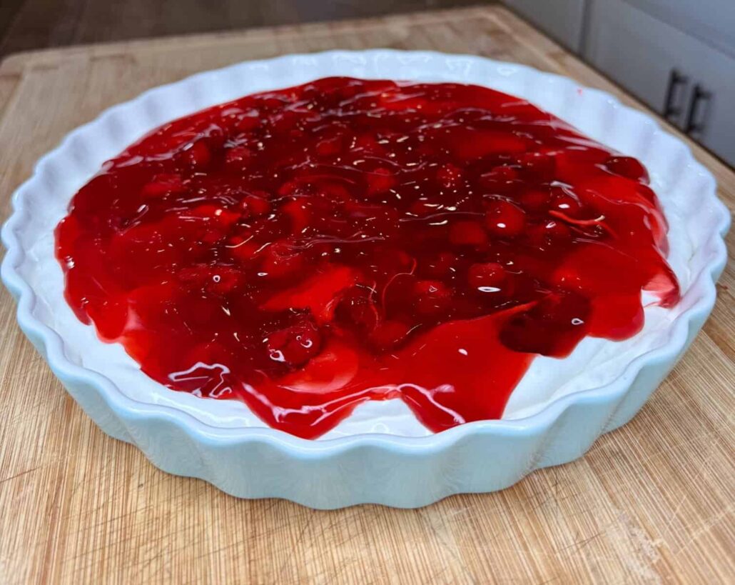 A ceramic pie dish filled with dessert topped with bright red strawberry or cherry filling, perfect for The Ultimate Easter Menu (Mediterranean & American), sits on a wooden surface in a cozy kitchen setting.