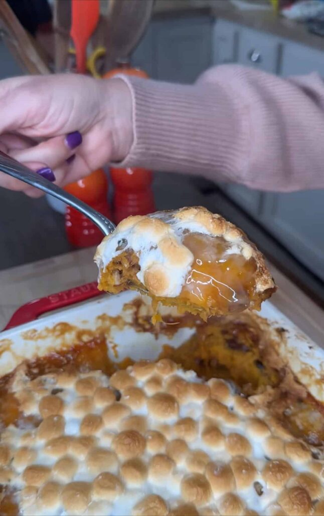 A hand holds a spoonful of sweet potato casserole topped with toasted marshmallows above a baking dish filled with the dessert in a kitchen setting.
