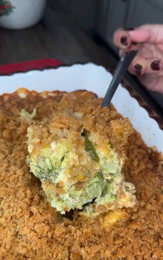 A hand with painted nails scoops out a serving of cheesy broccoli casserole topped with a golden, crispy breadcrumb crust from a white baking dish.
