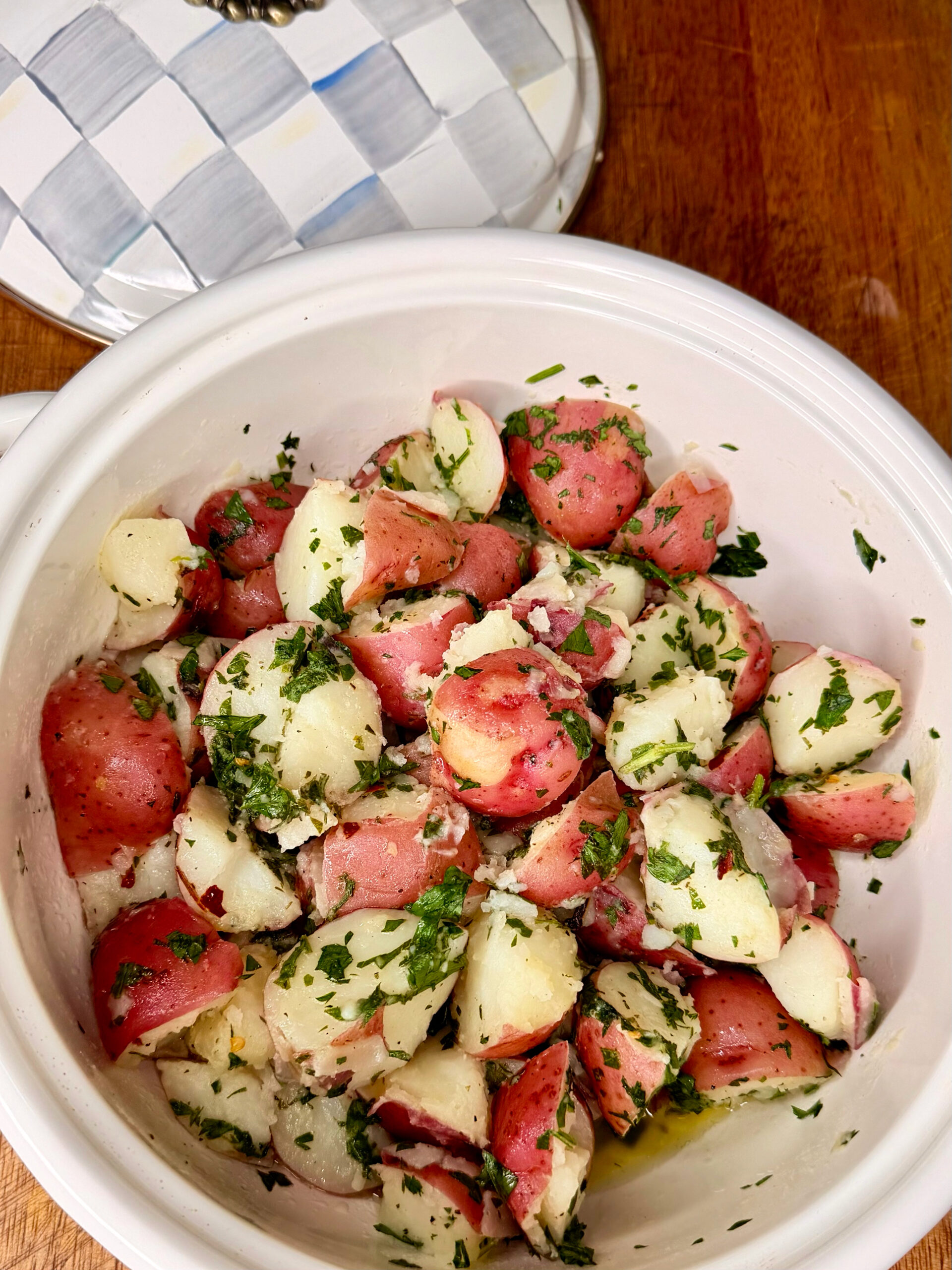 A white bowl filled with red-skinned potatoes cut into chunks, mixed with chopped parsley and herbs, sitting on a wooden surface next to a white dish with a checkered lid.